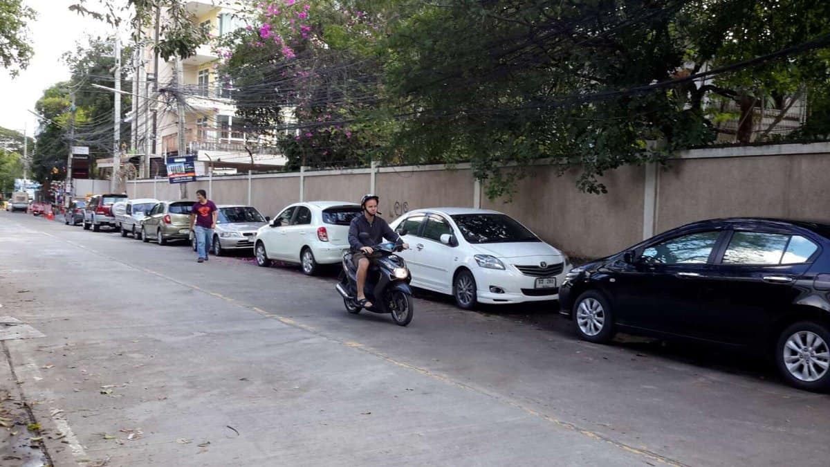 A clean street in Chiang Mai, Thailand, with parked cars along the sidewalk and a person riding a scooter in the center of the road. Trees and buildings line the street.