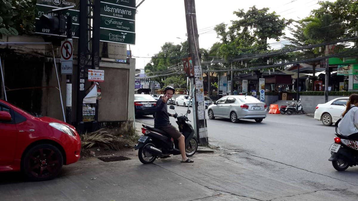 A person on a scooter gesturing with a peace sign on a busy street in Chiang Mai, Thailand. There are several cars and scooters on the road, and signs and foliage in the background.