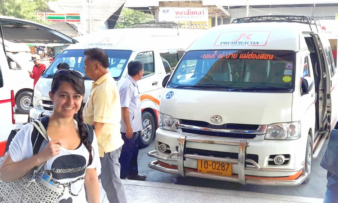 A smiling person is standing at a transport station in Chiang Mai, Thailand. Behind them are several white passenger vans, two of which have visible signage indicating they are part of the Prempracha Transport company. Two men are visible near the vans, presumably boarding or organizing travel arrangements.