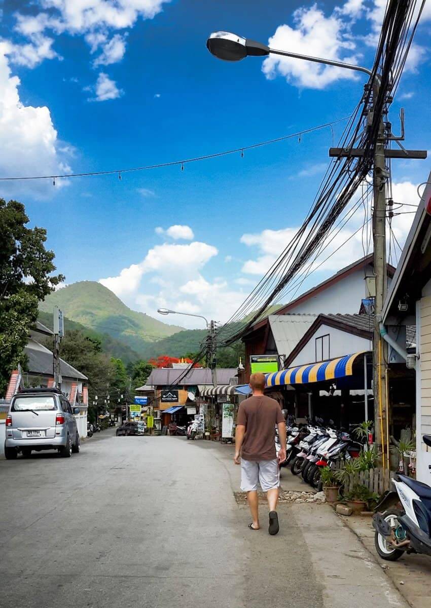 A man walking down a street in Pai, Thailand, with a scenic mountain landscape in the background.