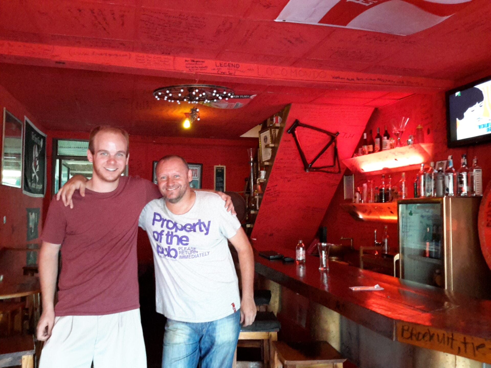 Two men posing for a photo inside the Hub Pub in Chiang Khong, Thailand. One of the men is Alan Bate, who holds the Guinness World Record for fastest circumnavigation of the globe by bicycle. The pub walls are covered with messages and signatures.