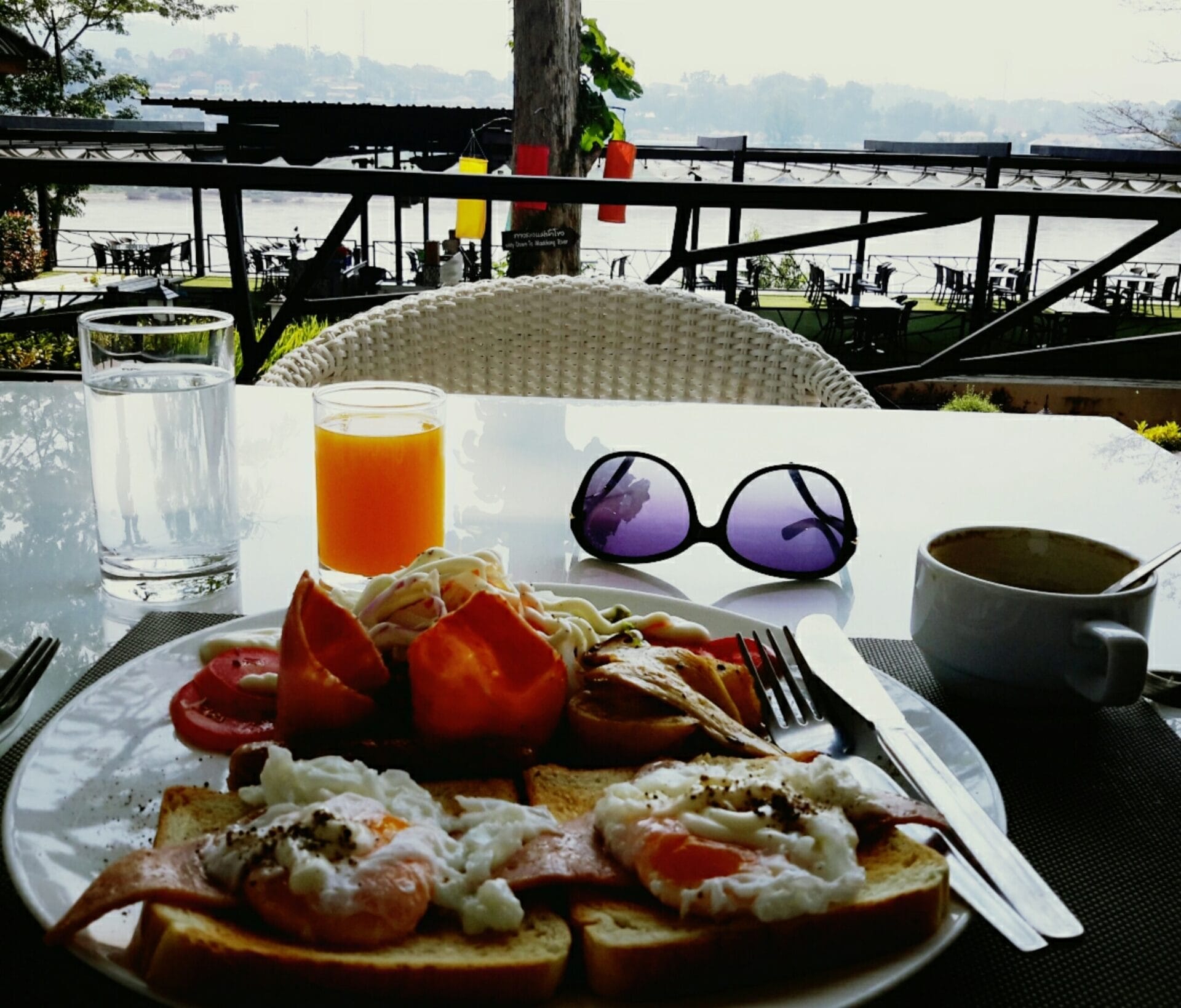 Breakfast at Chiangkhong Teak Garden Riverfront Onsen Hotel in Chiang Khong, Thailand. A plate of toast with eggs and ham, a glass of water, a glass of orange juice, a bowl of soup, and a pair of sunglasses on a table overlooking a river view.