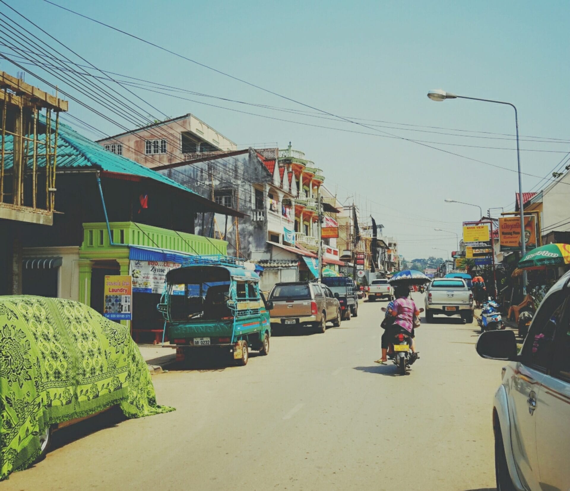 A busy street in Huay Xai, Laos, with various vehicles including trucks, a motorcycle, and local tuk-tuks. Numerous buildings, shops, and houses line both sides of the street, and the sky is clear.