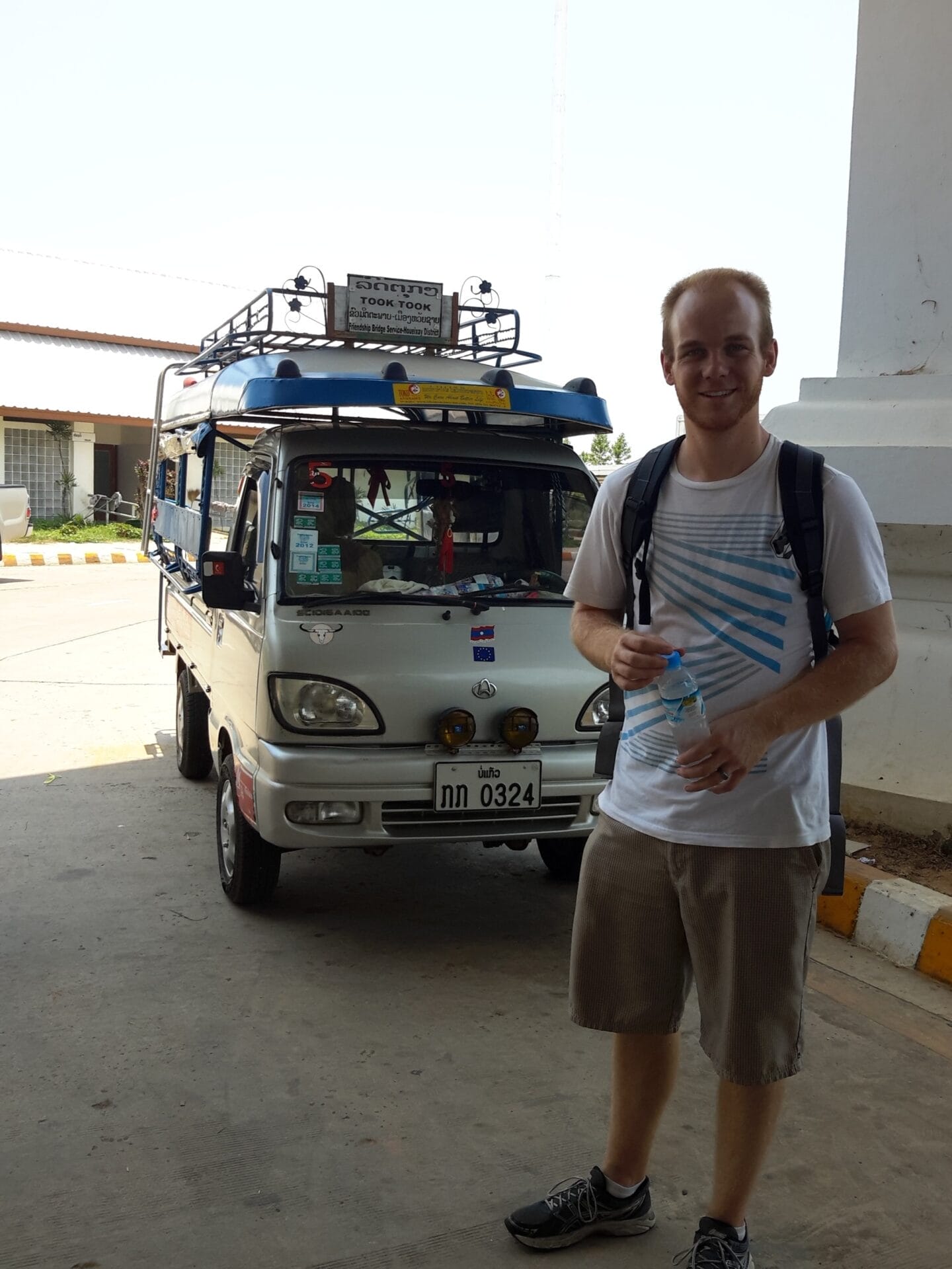 Man standing in front of a tuk-tuk in Huay Xai, Laos, holding a water bottle and wearing a backpack.