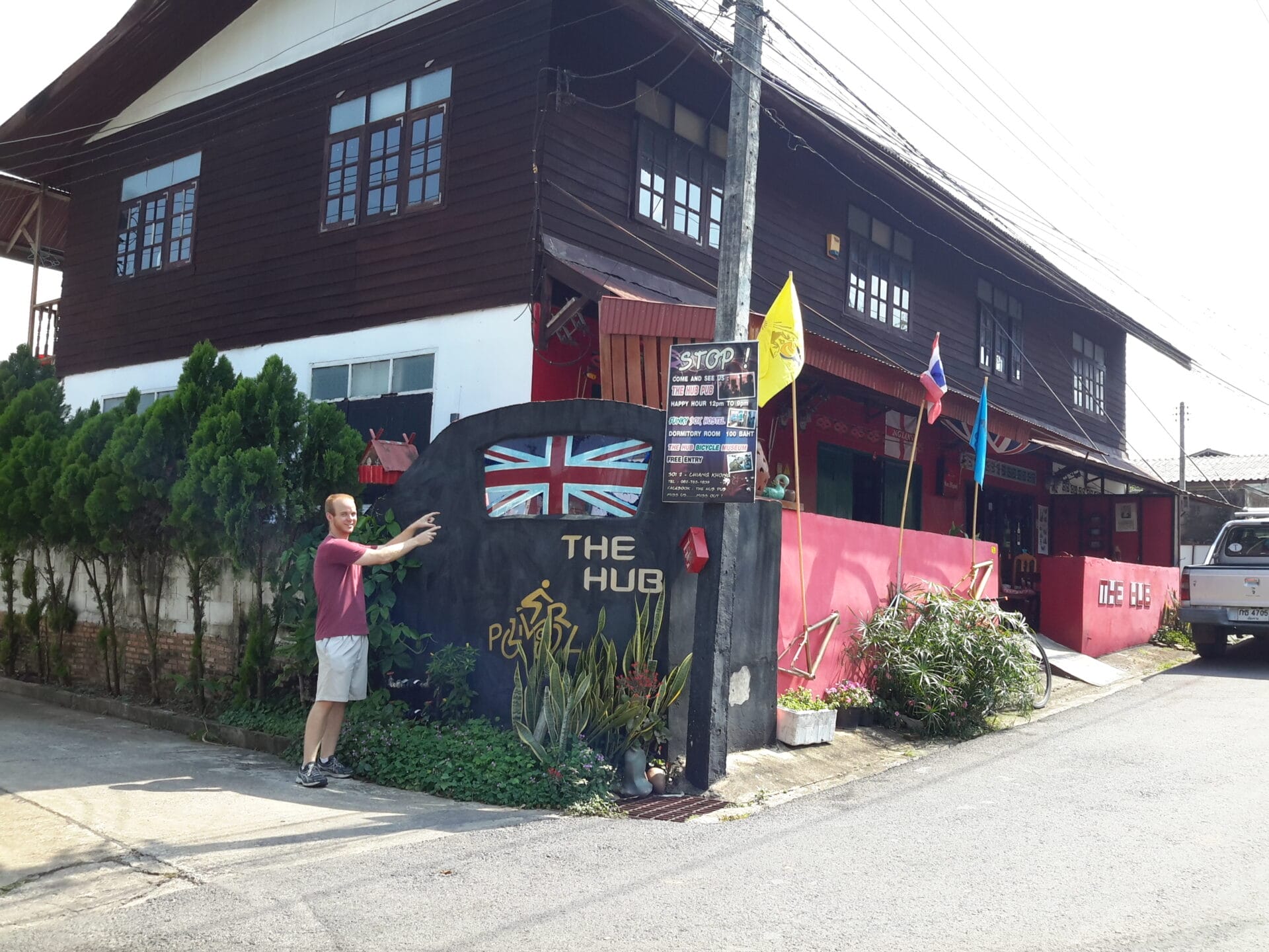 A person standing in front of The Hub Pub in Chiang Khong, Thailand, pointing at the establishment's sign which features a British flag.