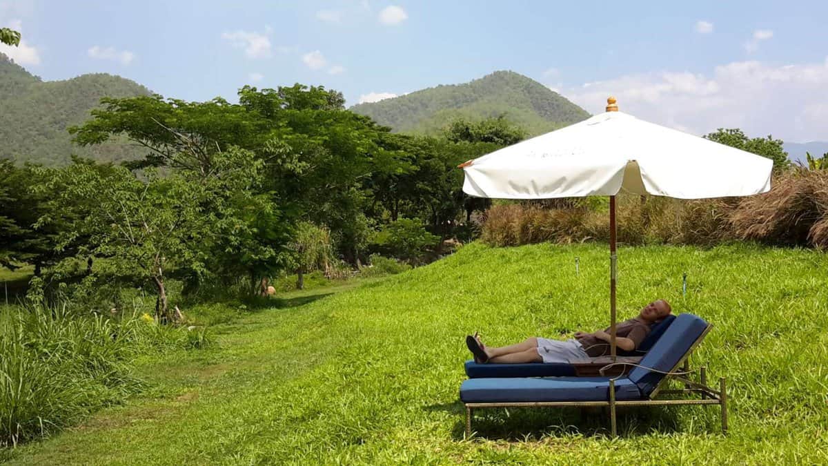 A person relaxing on a lounge chair under a large umbrella in a green field, surrounded by trees and hills in Pai, Thailand.