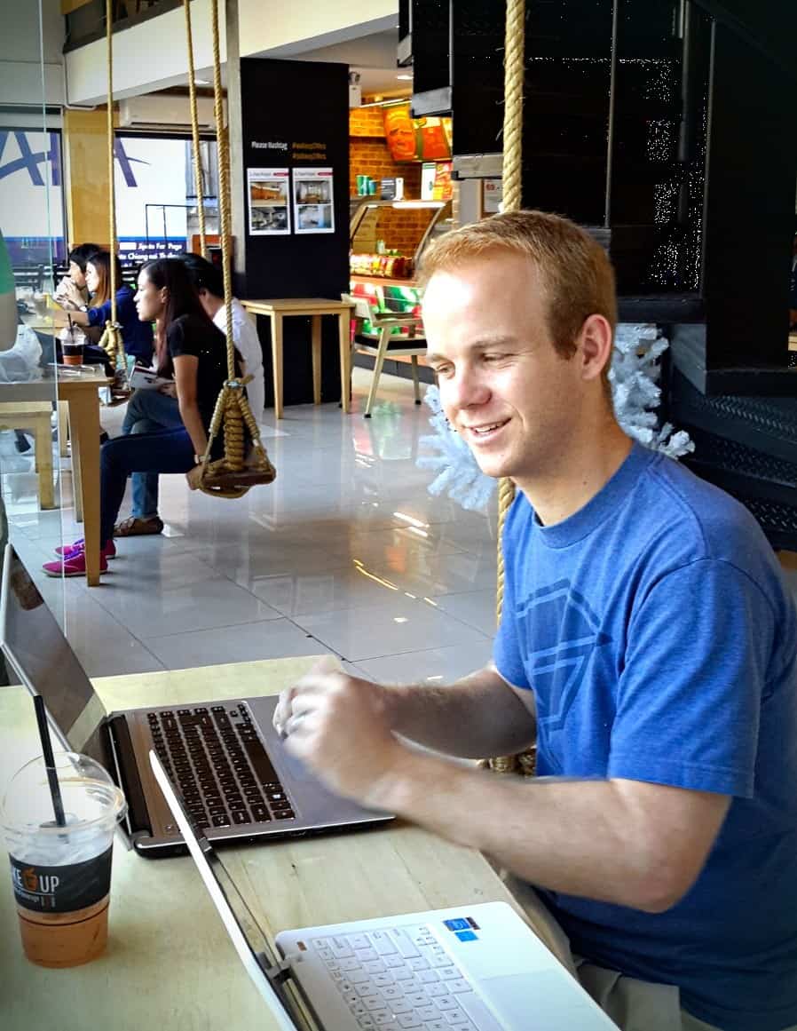 A person smiling while working on a laptop at a cafe in Chiang Mai, Thailand. Other patrons are in the background, some sitting at tables and others working on laptops. The cafe features indoor swings as seating.