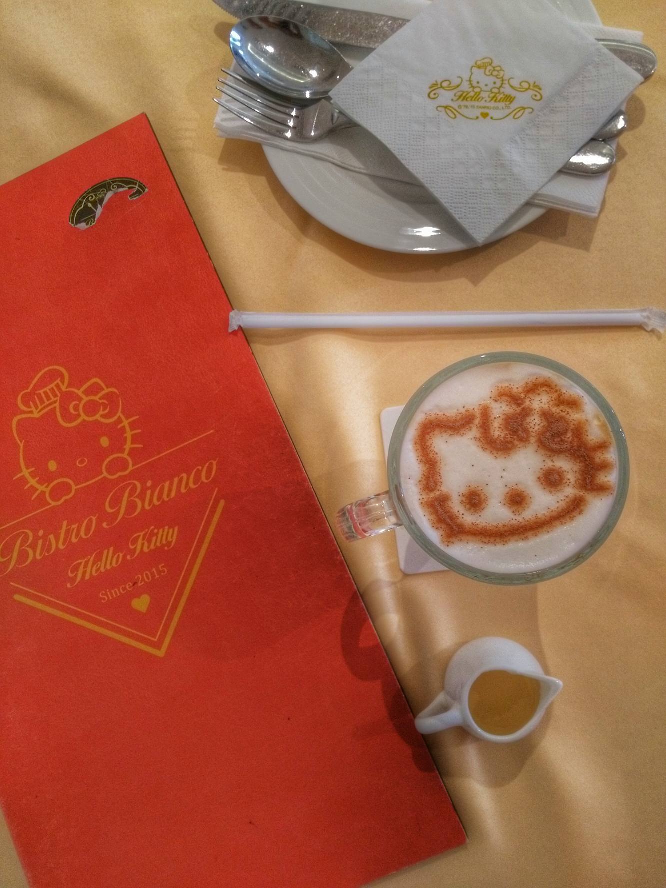 A table at Bistro Bianco Hello Kitty themed café on Nanjing Road in Shanghai, China, featuring a red menu with a Hello Kitty logo, a cup of coffee with a Hello Kitty design on the foam, white napkins with Hello Kitty logos, and a plate with cutlery.