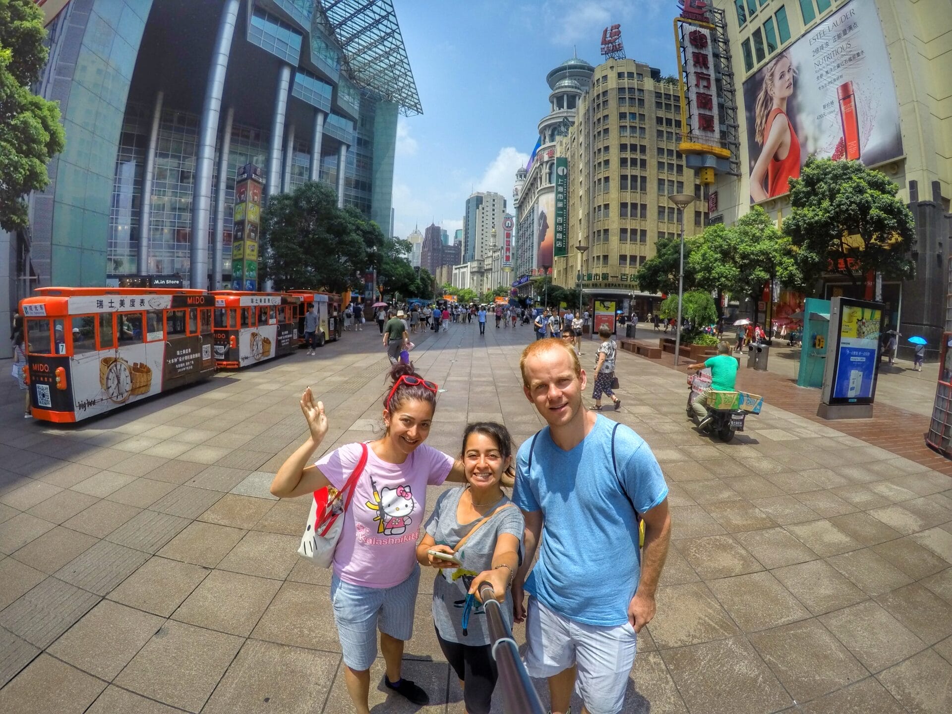 Three people posing for a selfie on Nanjing Road in Shanghai, China, with a busy street, modern buildings, and an orange bus in the background on a sunny day.