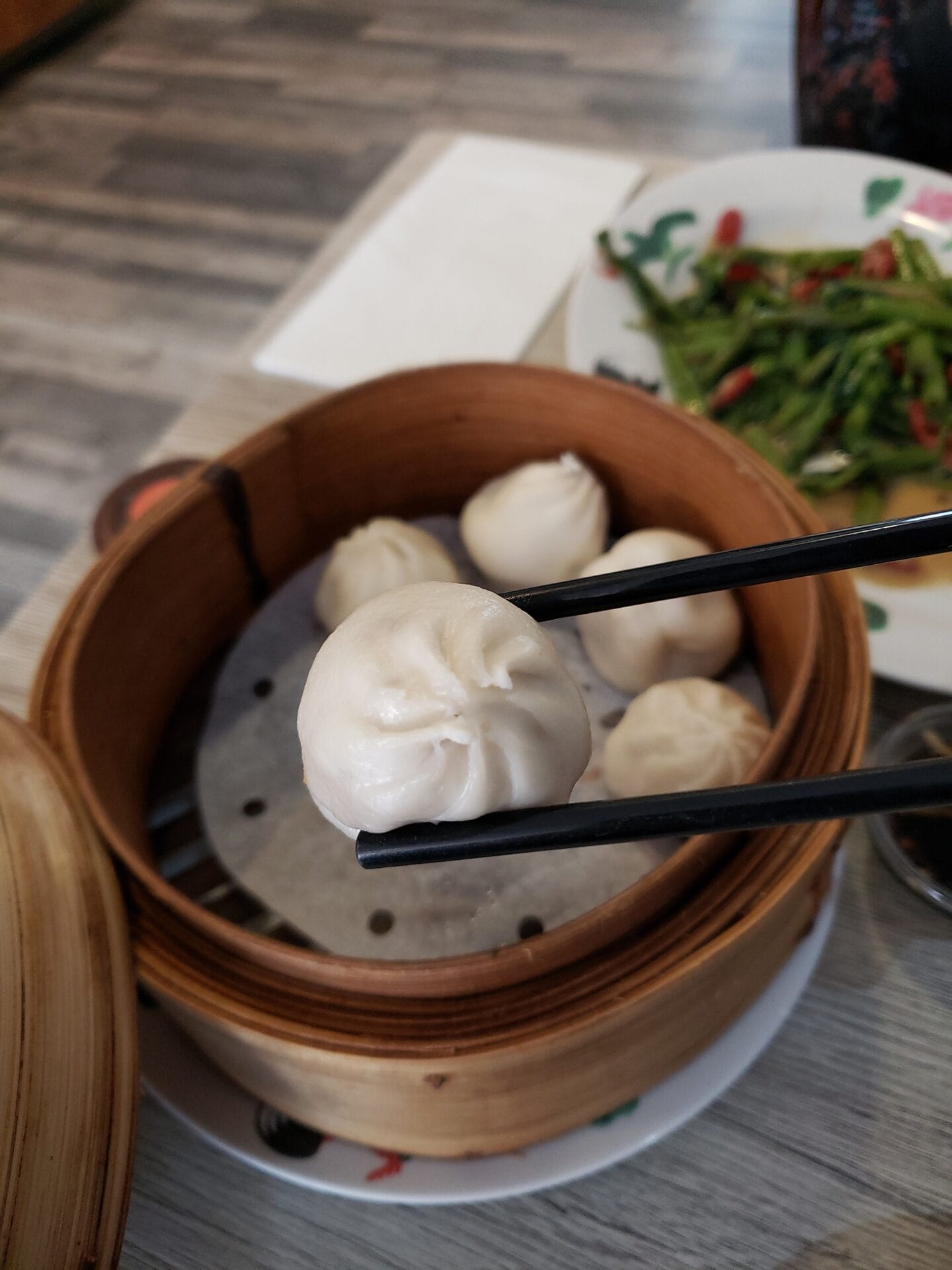 A close-up of a pair of chopsticks holding a steamed dumpling over a bamboo steamer filled with more dumplings, with a plate of greens in the background. The scene is set in a restaurant in Shanghai, China.