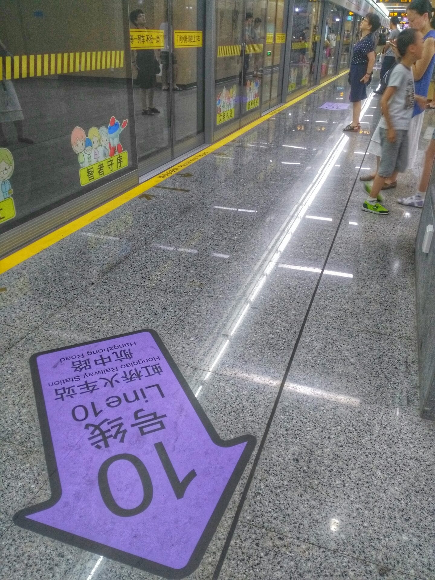People waiting on the platform at a Shanghai Metro station in Shanghai, China. A purple arrow on the floor indicates "Line 10" and the direction to board. The platform doors have cartoon decorations and safety markings in yellow.