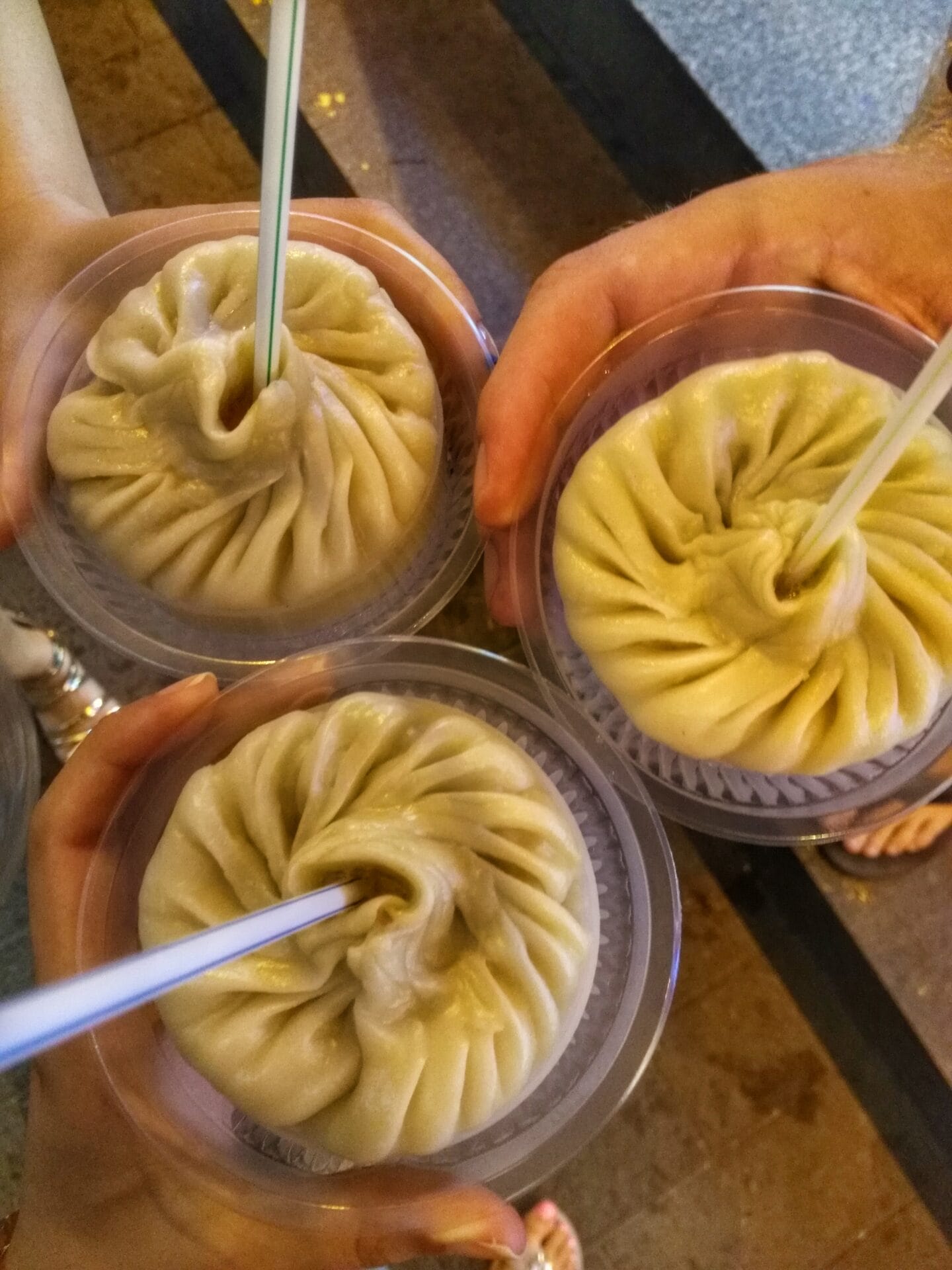 Three bowls of soup dumplings with straws held by three people at Yu Garden in Shanghai, China.