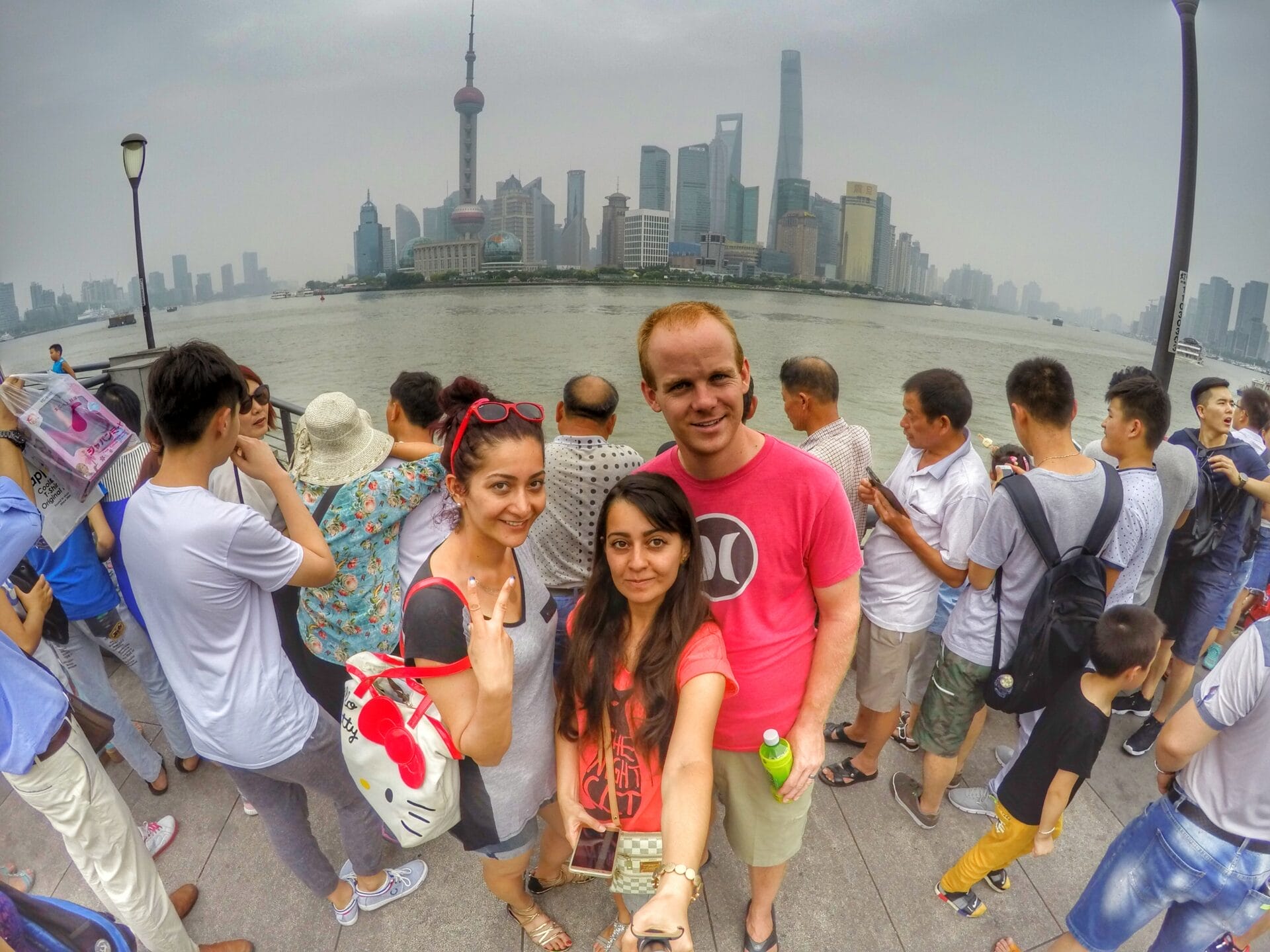 A group of people taking a selfie with the skyline of The Bund in Shanghai, China in the background. They are standing on a crowded promenade with the Oriental Pearl Tower and modern skyscrapers visible across the river.