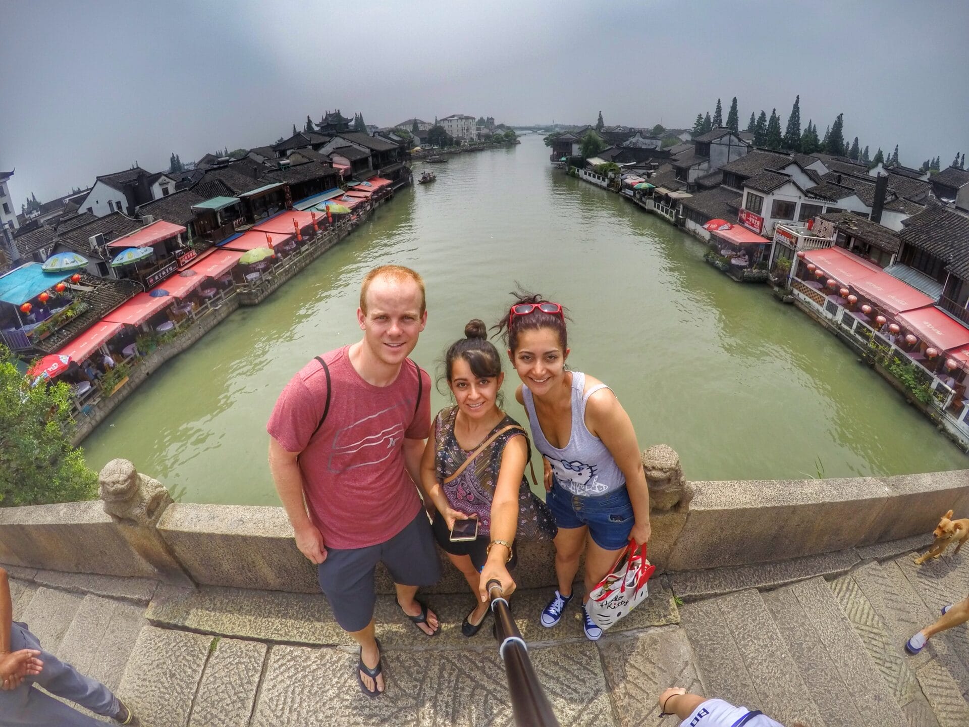 Three people taking a selfie on a bridge in Zhujiajiao, a water town in Shanghai, China, with traditional buildings and a canal in the background.