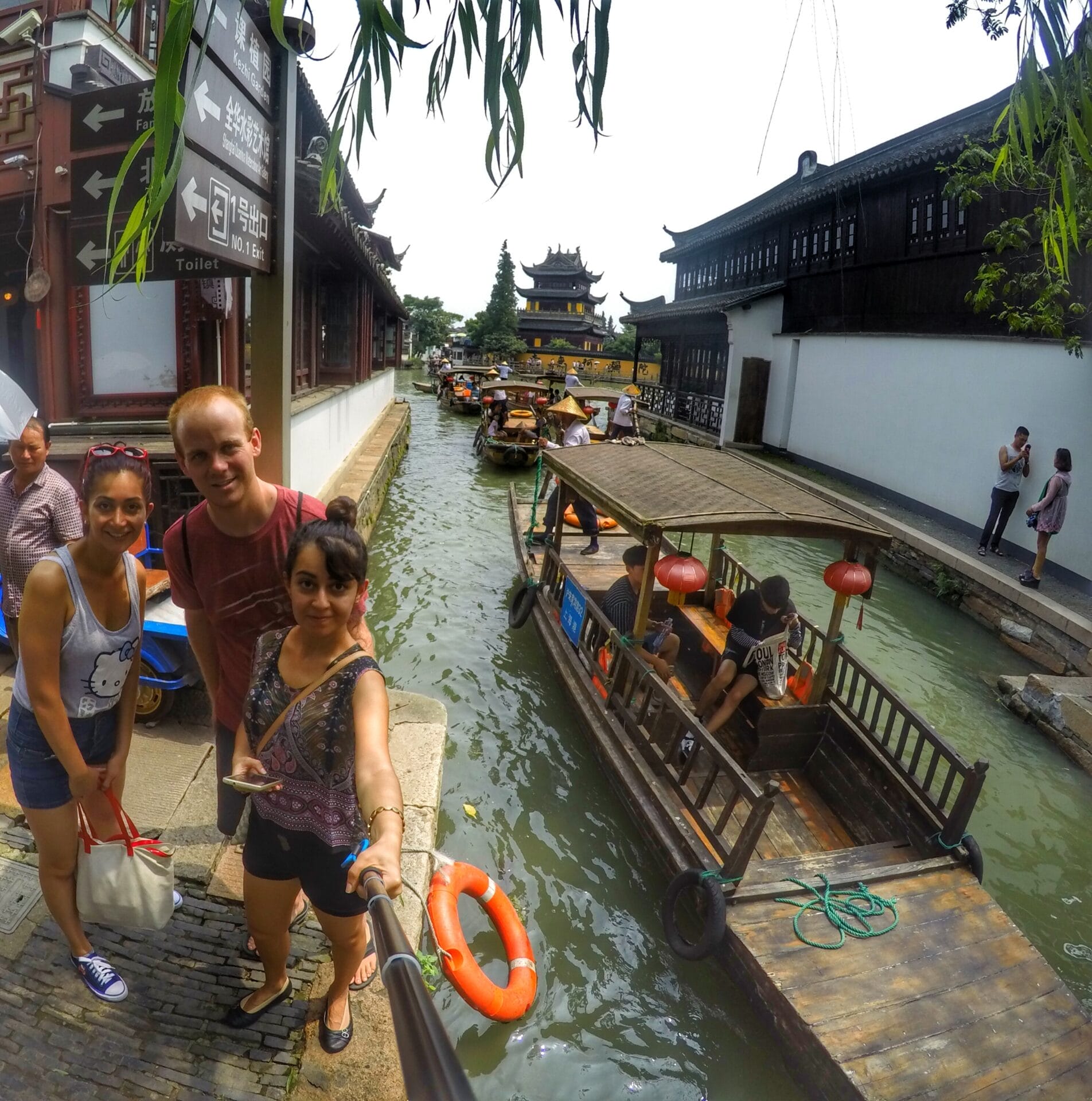 Tourists taking a selfie near a canal in Zhujiajiao, Shanghai, China, with traditional boats and historic buildings in the background.