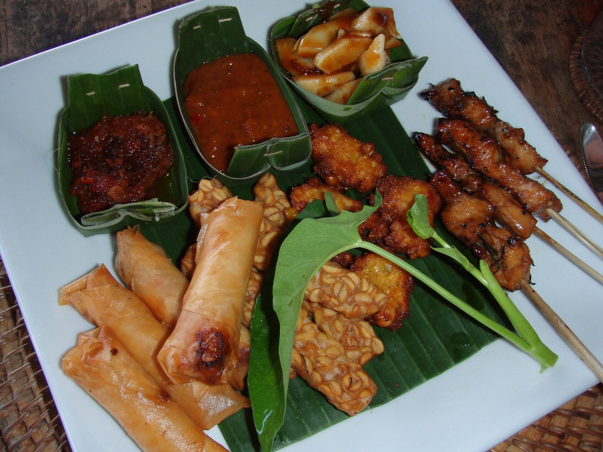 A plate of diverse Indonesian dishes served in Bali, Indonesia. The plate includes skewers of grilled meat, spring rolls, tempeh, fritters, and a selection of dipping sauces, arranged on banana leaves.