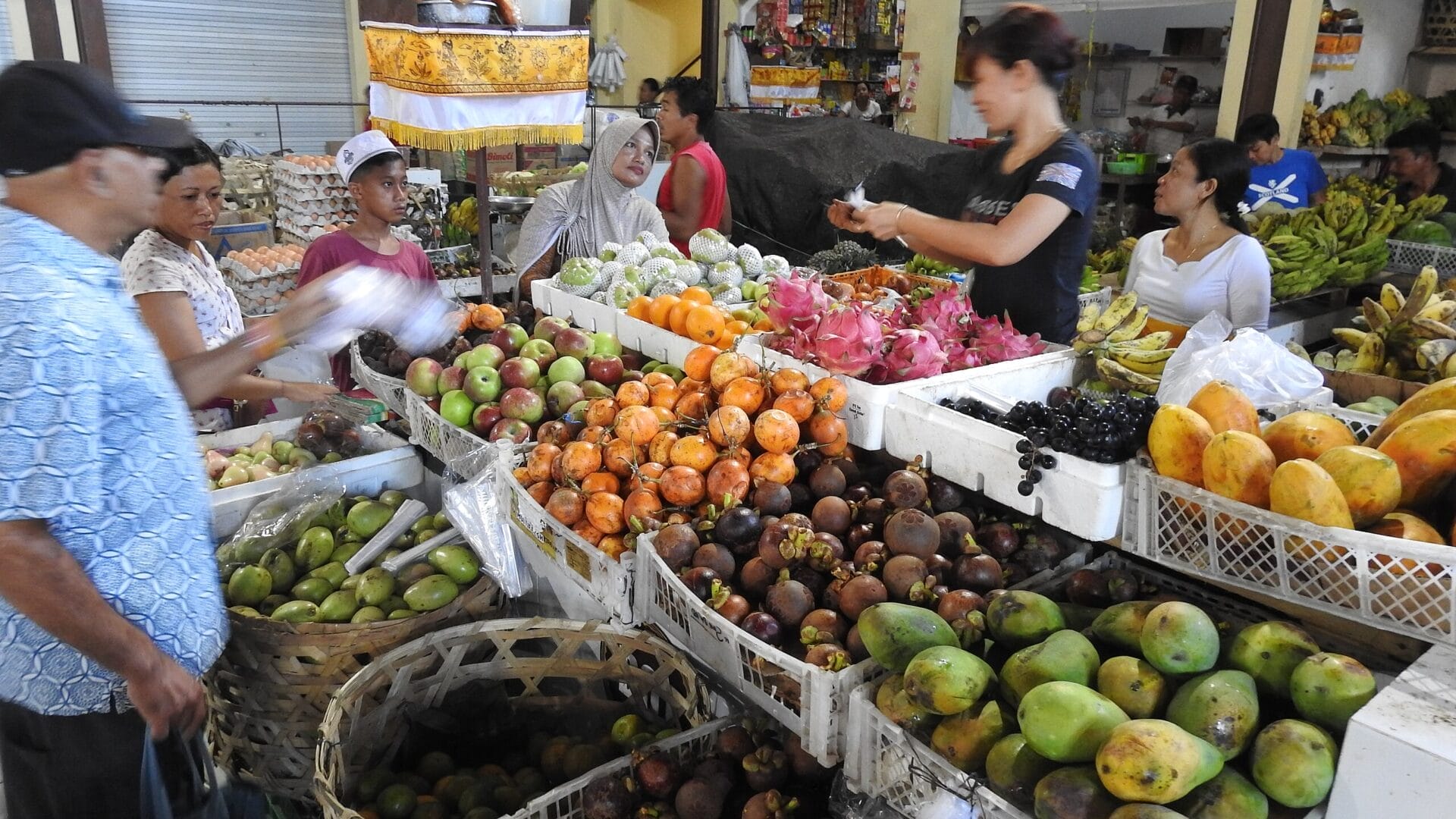 A bustling local market in Bali, Indonesia, with an array of fresh fruits including mangoes, apples, dragon fruit, and bananas displayed at several vendor stalls. Several people are shopping and interacting with the vendors.