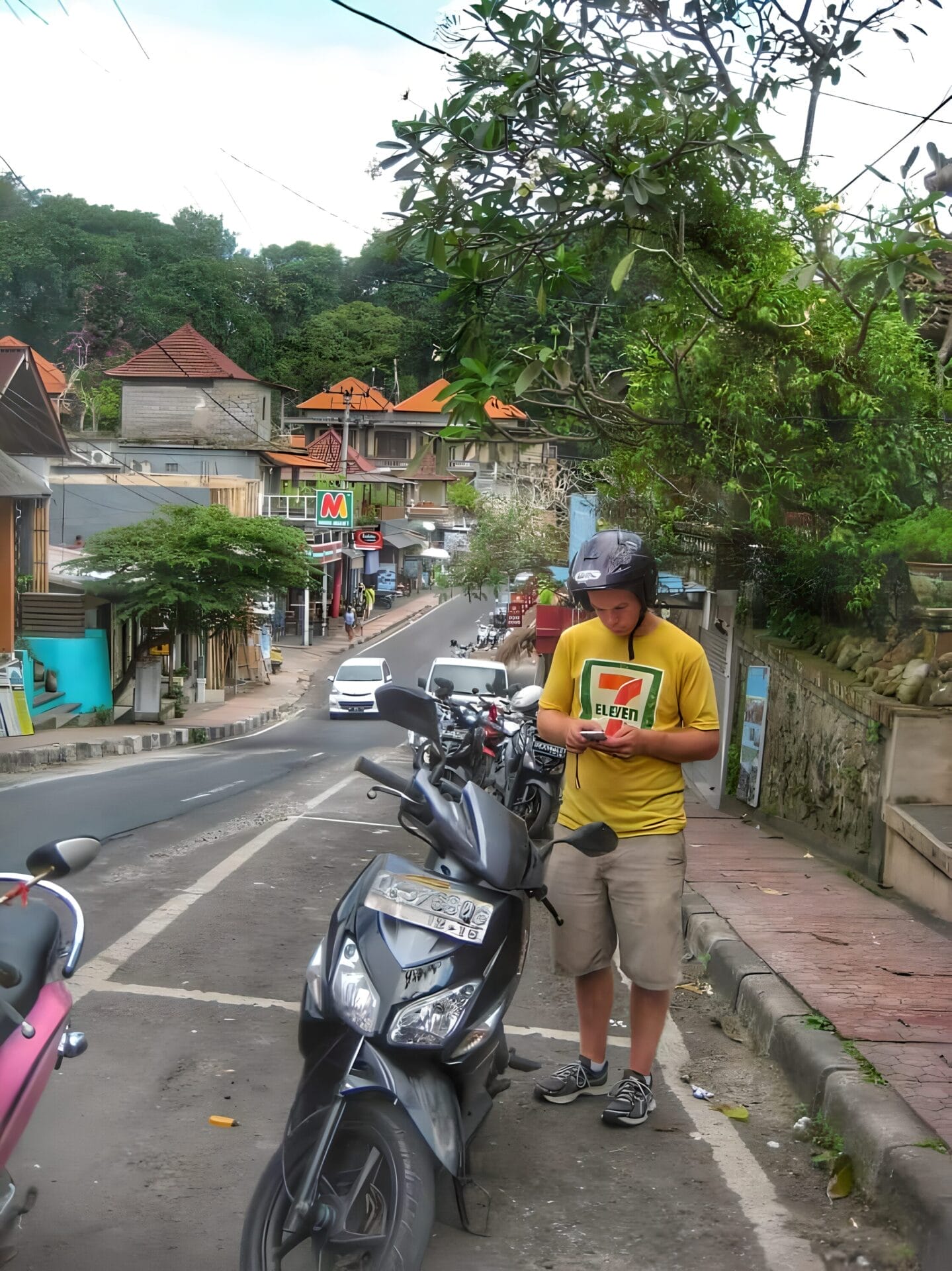 A person stands next to a parked scooter on a street in Ubud, Bali. Various shops line the street, and tropical foliage surrounds the area. The person is wearing a yellow t-shirt with a logo, shorts, and has a helmet on.