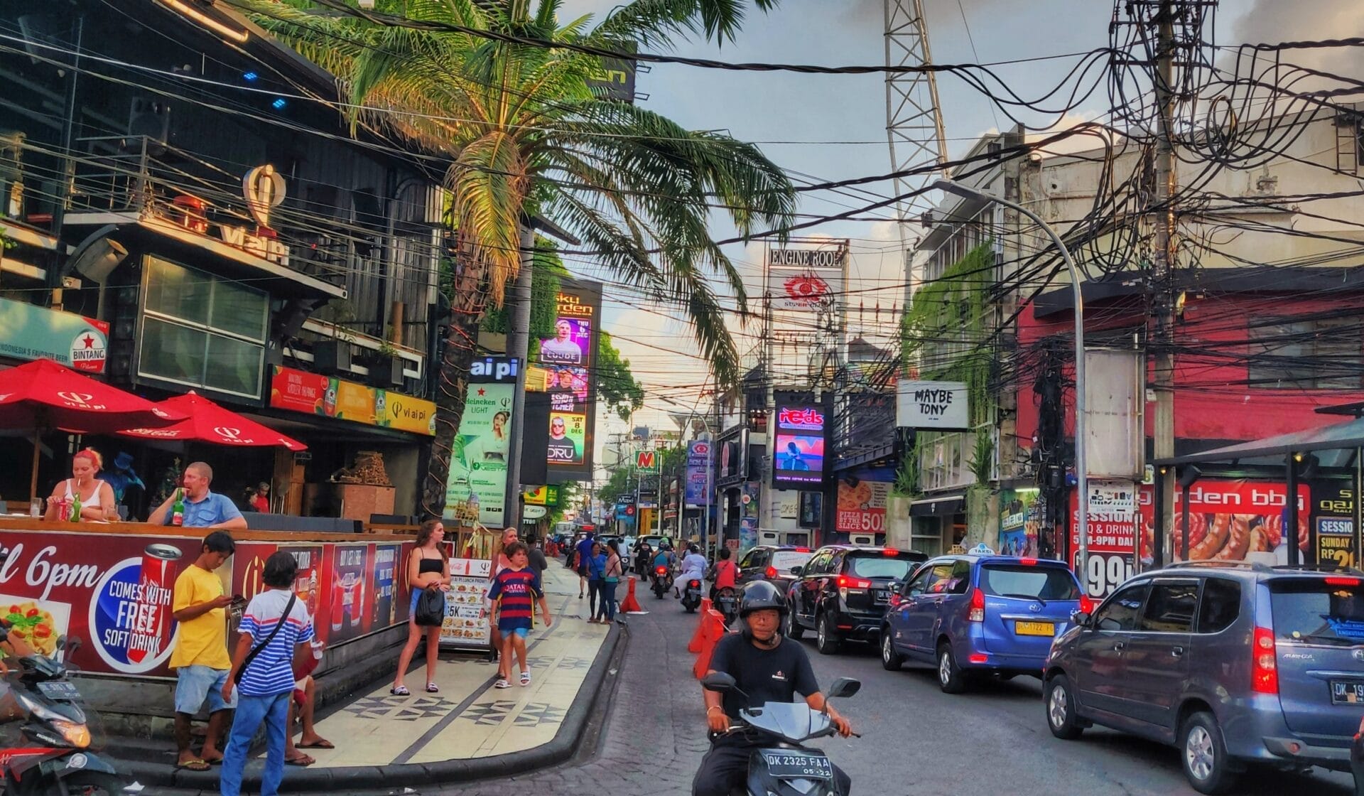 A busy street in Bali, Indonesia, lined with shops, restaurants, and palm trees. People are walking on the sidewalk and a man riding a scooter is heading down the street. 