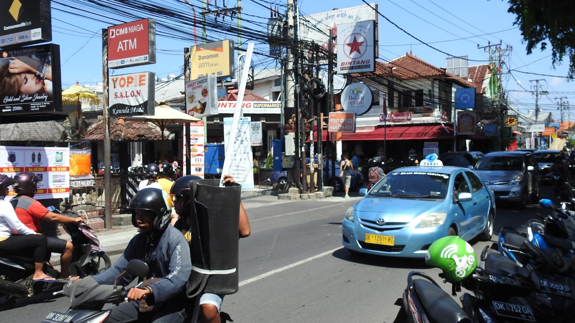 Busy street scene in Bali, Indonesia with motorcycles, a blue taxi, and various signs for local businesses.