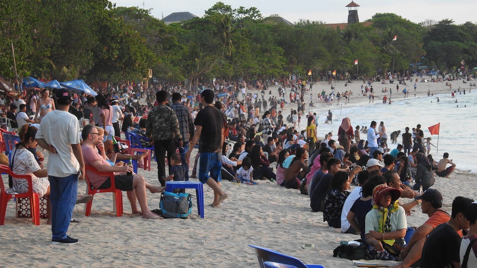 Crowded beach in Bali, Indonesia, with people sitting on colorful plastic chairs and the sandy shore.