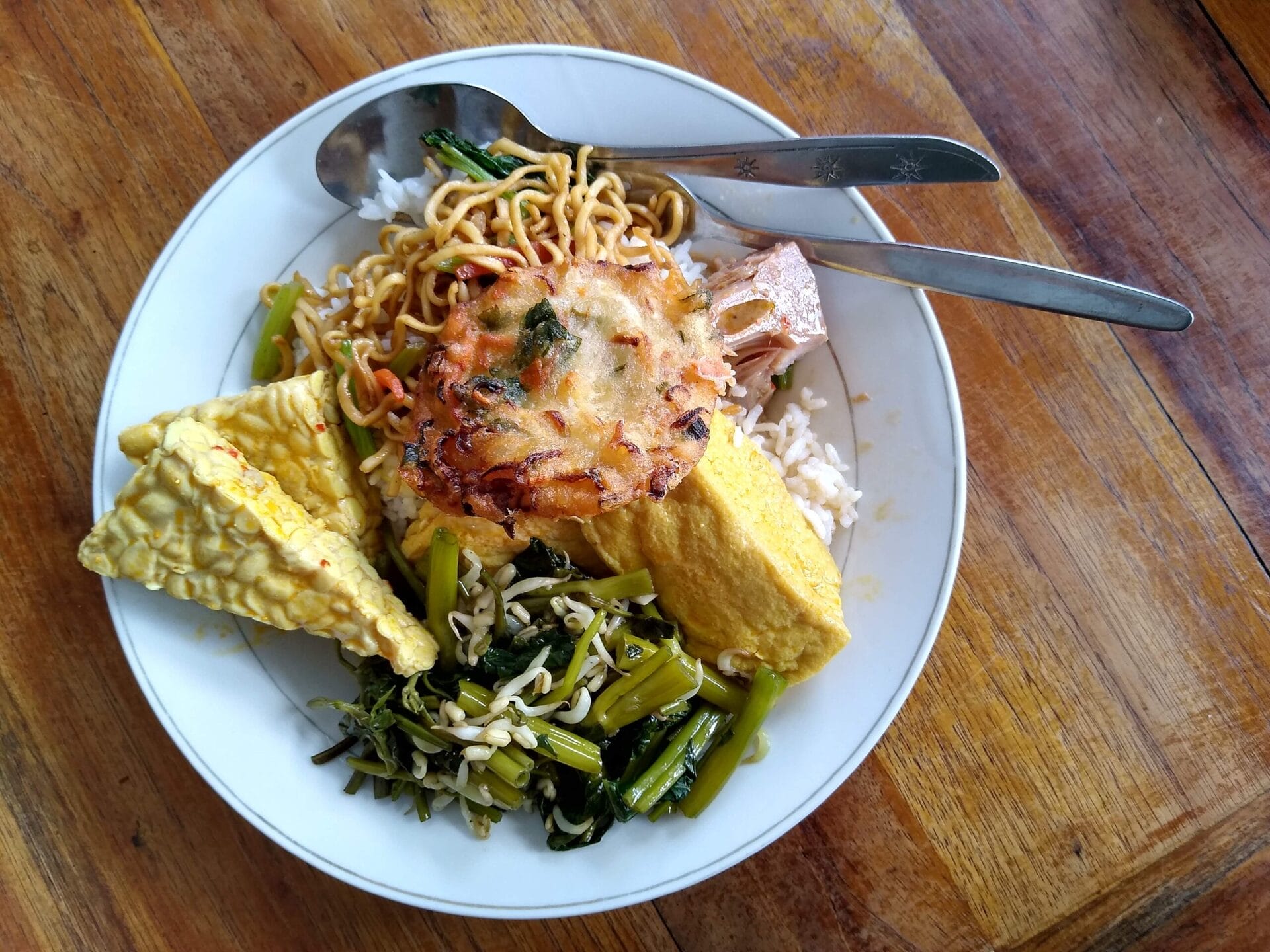A plate of Nasi Campur, a traditional Indonesian dish, featuring rice, noodles, vegetables, tofu, and a savory fritter, served on a wooden table in Bali, Indonesia.
