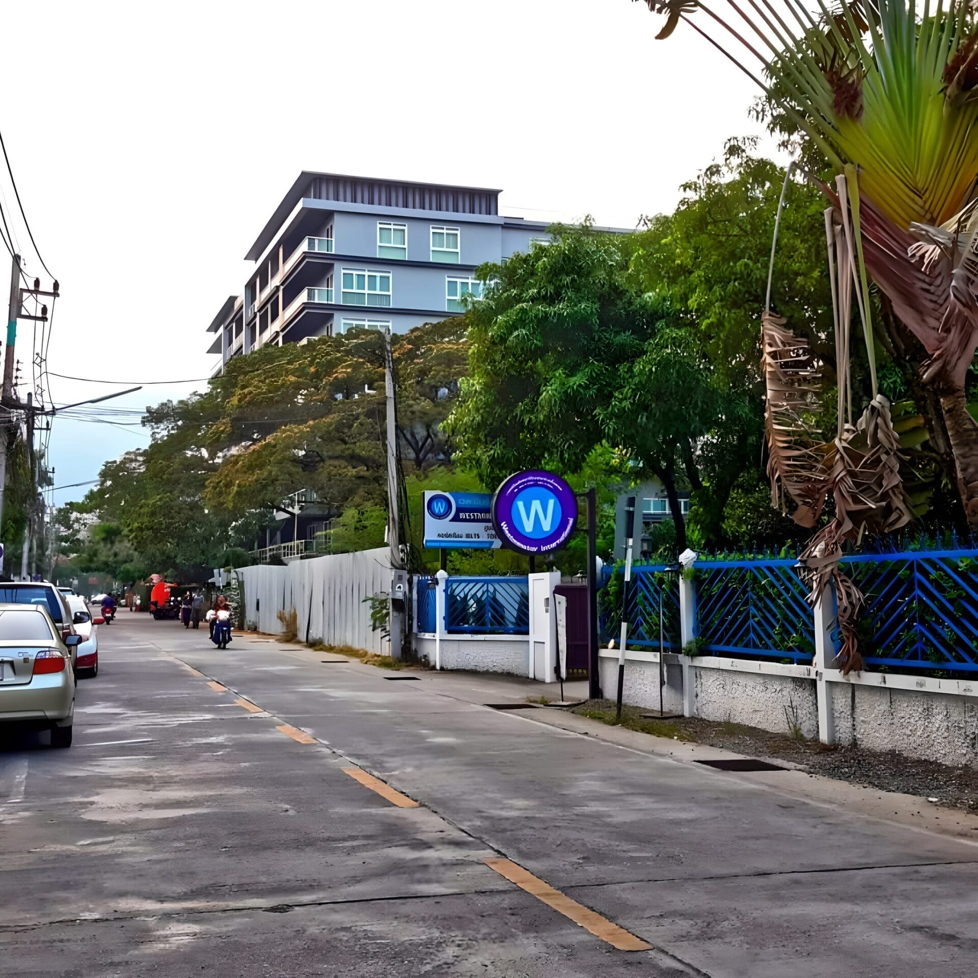 Street view near Rawee Waree Condominium in Chiang Mai, Thailand, showing parked cars, motorbike riders, and a fence with signage.