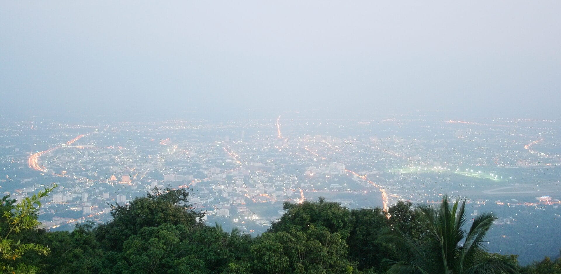 A hazy view of Chiang Mai, Thailand during the burning season. City lights are visible through the thick haze, with tree-covered hills in the foreground.