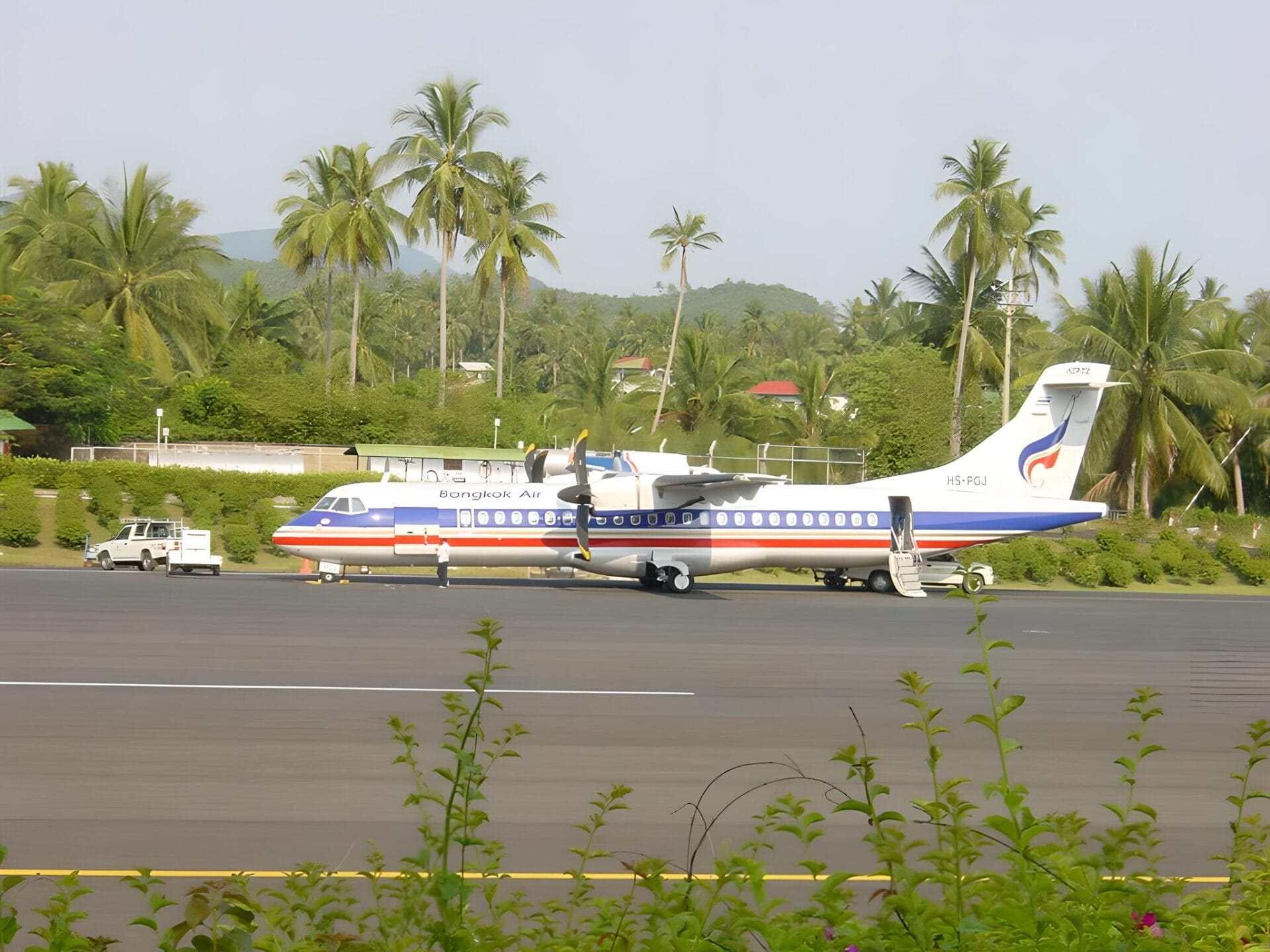 A Bangkok Airways plane parked on the tarmac at Koh Samui International Airport in Thailand, surrounded by lush greenery and palm trees.