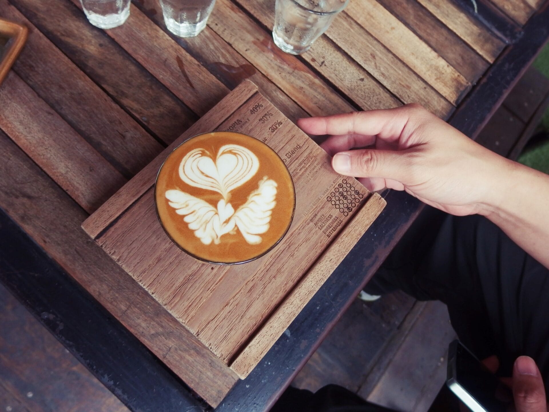 A hand reaches towards a beautifully crafted latte with intricate latte art resembling a heart and wings, served on a wooden tray at Ristr8to in Chiang Mai, Thailand. The tray has details about the coffee written on it. 