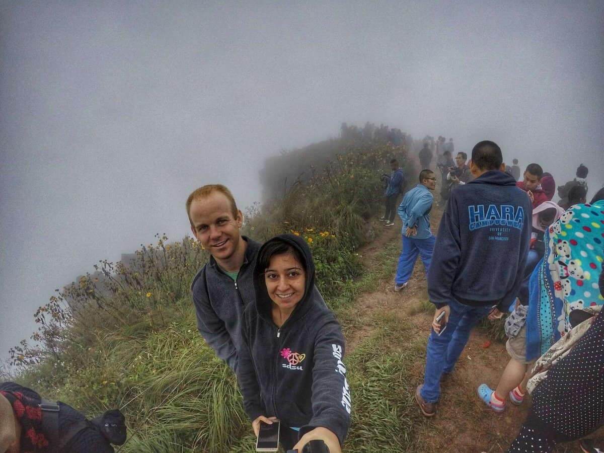 A group of tourists standing on the misty peak of Phu Chi Fa, near Chiang Rai, Thailand. Two individuals are taking a selfie at the front, capturing the foggy landscape in the background.