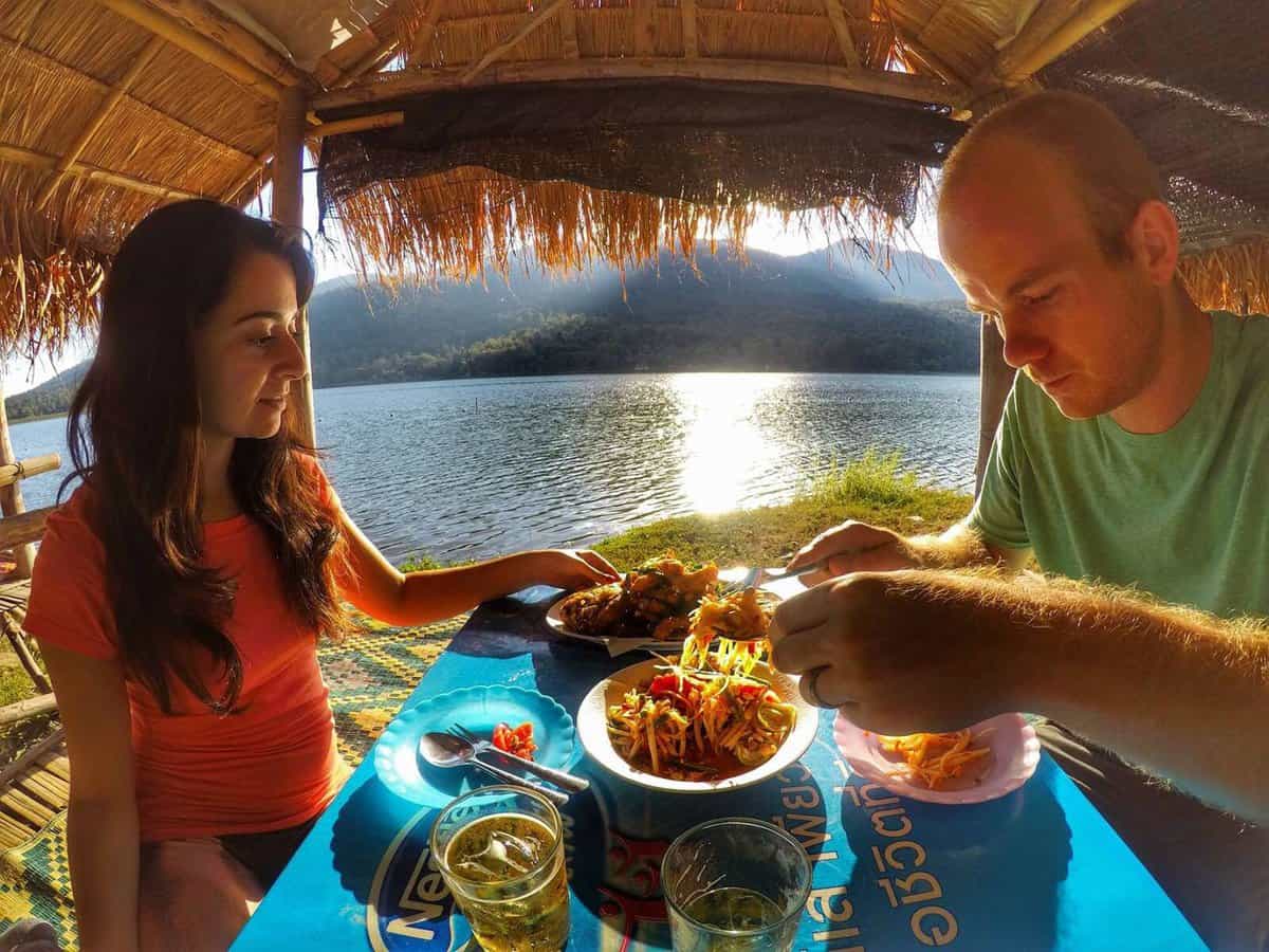 A couple dining in a bamboo hut at Huai Tung Tao Lake in Chiang Mai, Thailand, with a scenic view of the lake and surrounding mountains in the background.