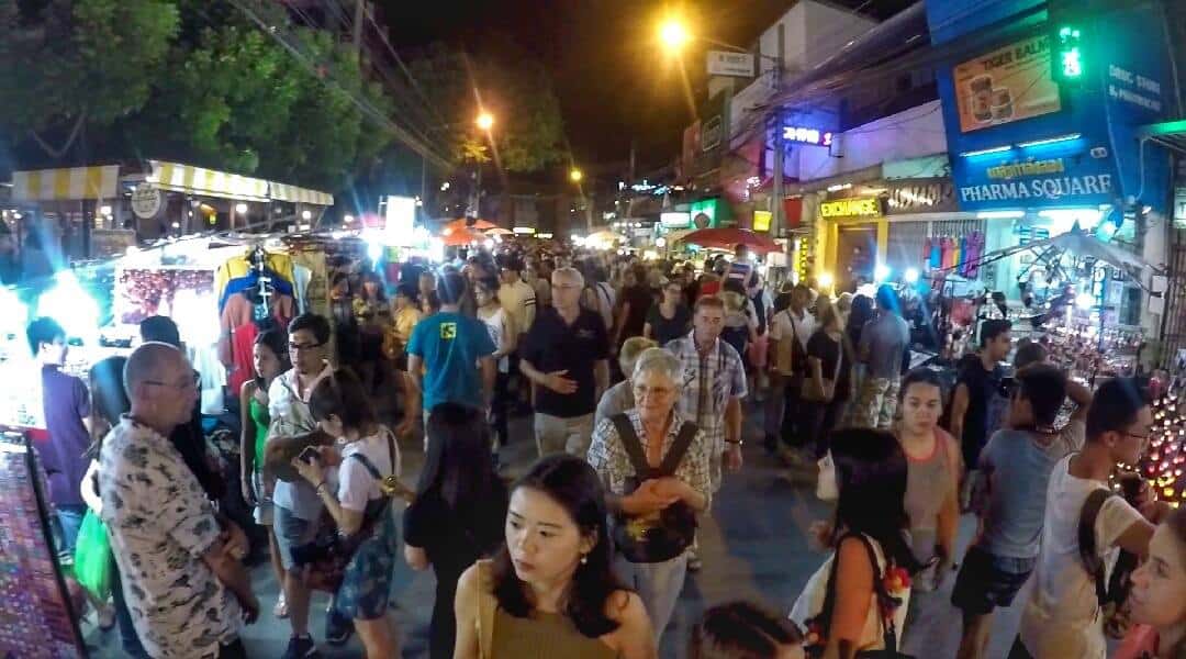 A bustling scene at the Chiang Mai Night Bazaar in Thailand, with crowds of people walking and exploring various market stalls and shops under the glowing streetlights at night.