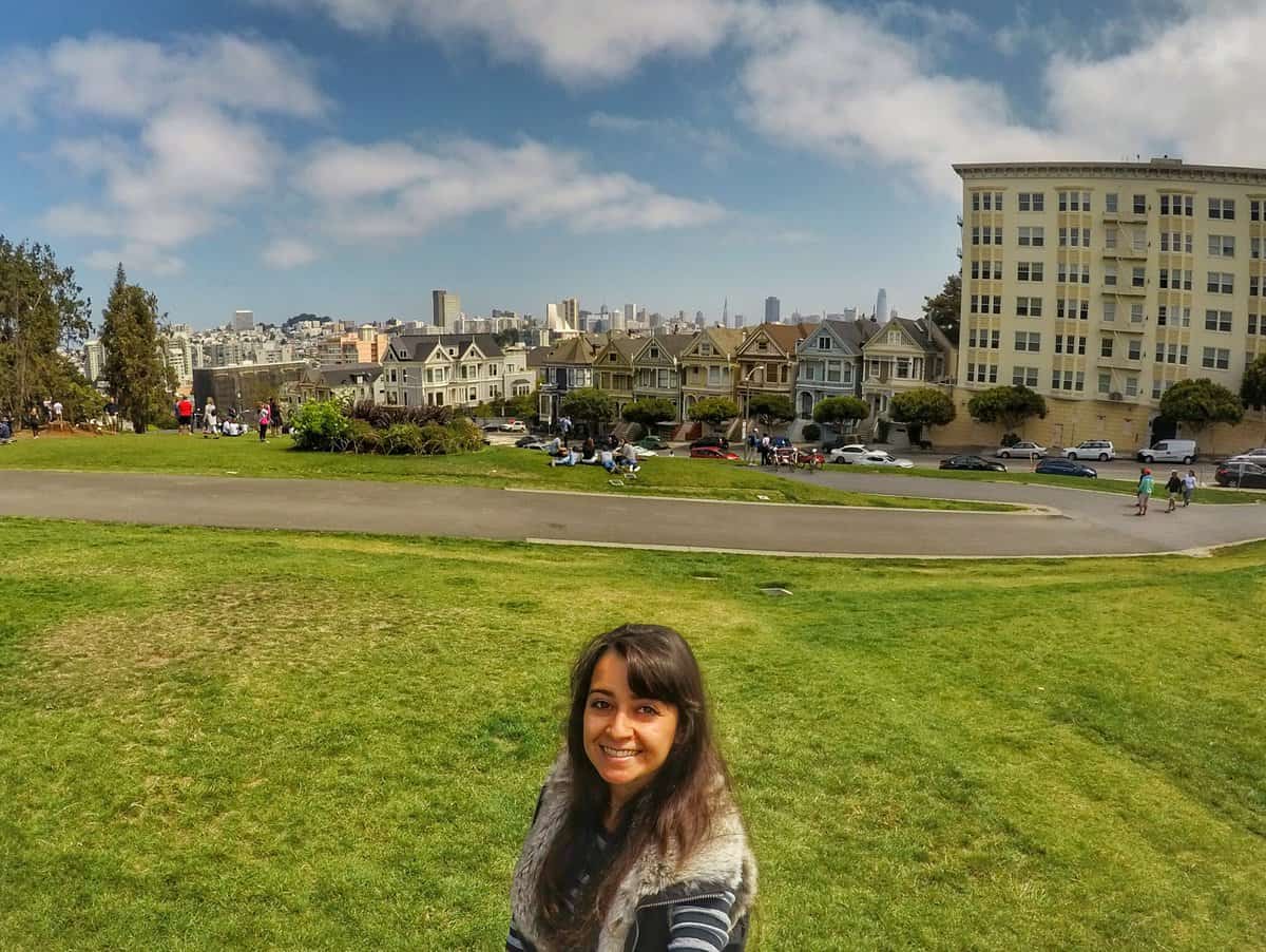 A woman is standing and smiling in a park with a green grassy area in San Francisco, with the iconic Painted Ladies and city skyline in the background.