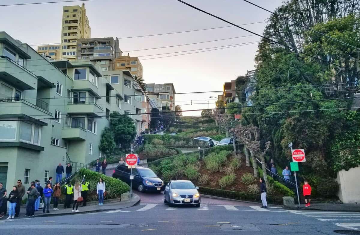 A view of Lombard Street in San Francisco with cars navigating the famous winding road, surrounded by residential buildings and lush greenery, as people watch from the sidewalks.