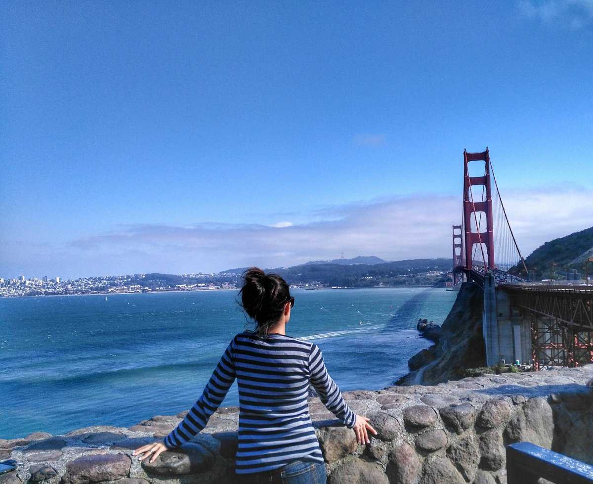 A person in a striped shirt stands at a stone railing overlooking the Golden Gate Bridge and San Francisco Bay on a clear day.