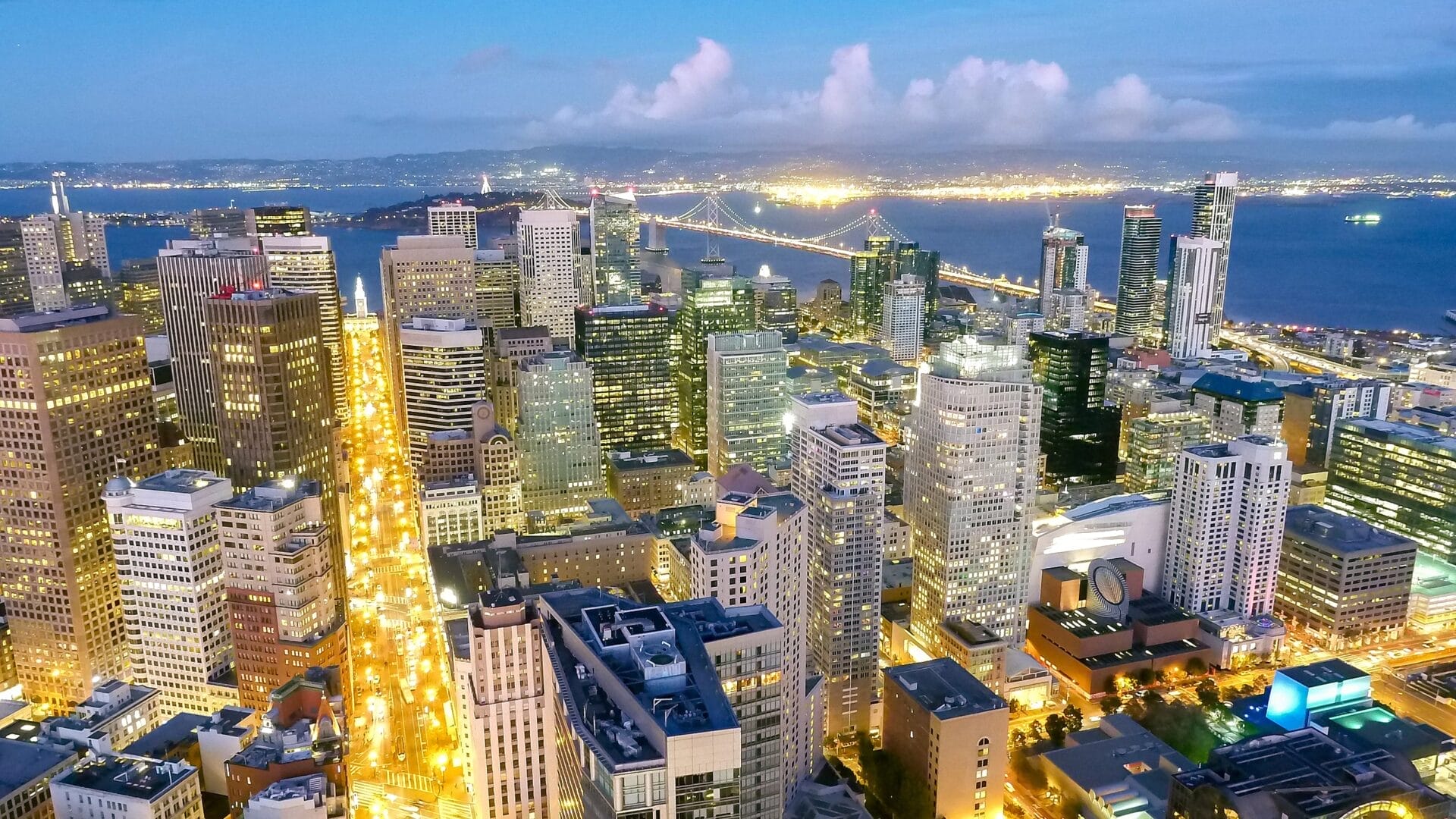 Aerial view of the Financial District in San Francisco at dusk, showcasing illuminated skyscrapers and the Bay Bridge in the background.