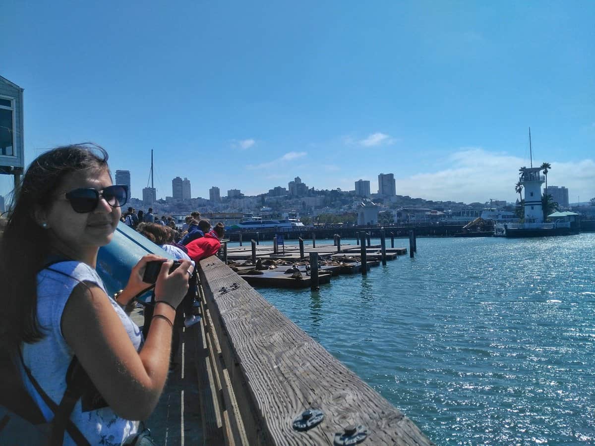 A woman with sunglasses and holding a camera, standing on a wooden pier at Fisherman's Wharf in San Francisco, overlooking the water and cityscape in the background.