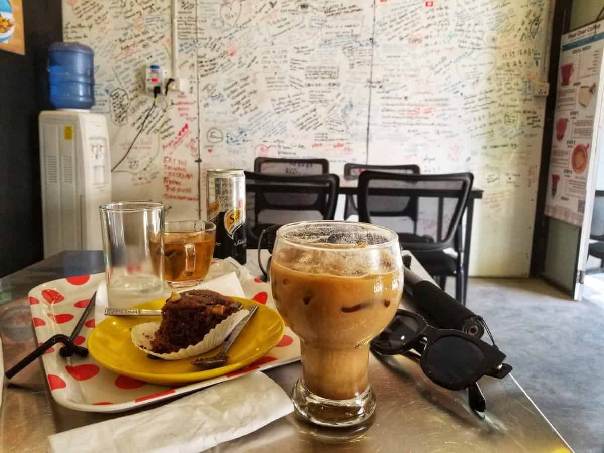 Bagan Travel guide and tips A cozy cafe in Nyaung-U, Bagan, Myanmar, displaying a table with a yellow plate holding a piece of cake, an iced coffee in a glass, a pair of sunglasses, an empty glass, and a can of soda. The cafe's walls are decorated with handwritten messages.