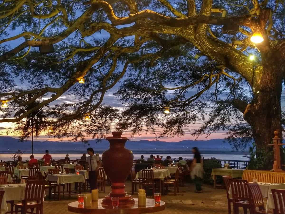 Myanmar Bagan Travel Guide Outdoor dining area at Thande Hotel in Old Bagan, Myanmar, beautifully lit under a large tree during sunset with the river and mountains in the background.