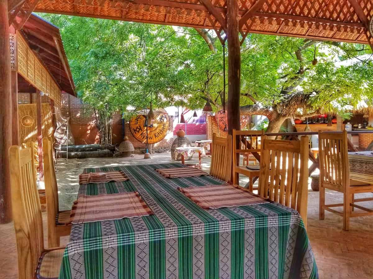 New Bagan restaurant Outdoor dining area at a restaurant in New Bagan, Myanmar, with wooden tables and chairs, colorful tablecloths, and lush trees providing shade.