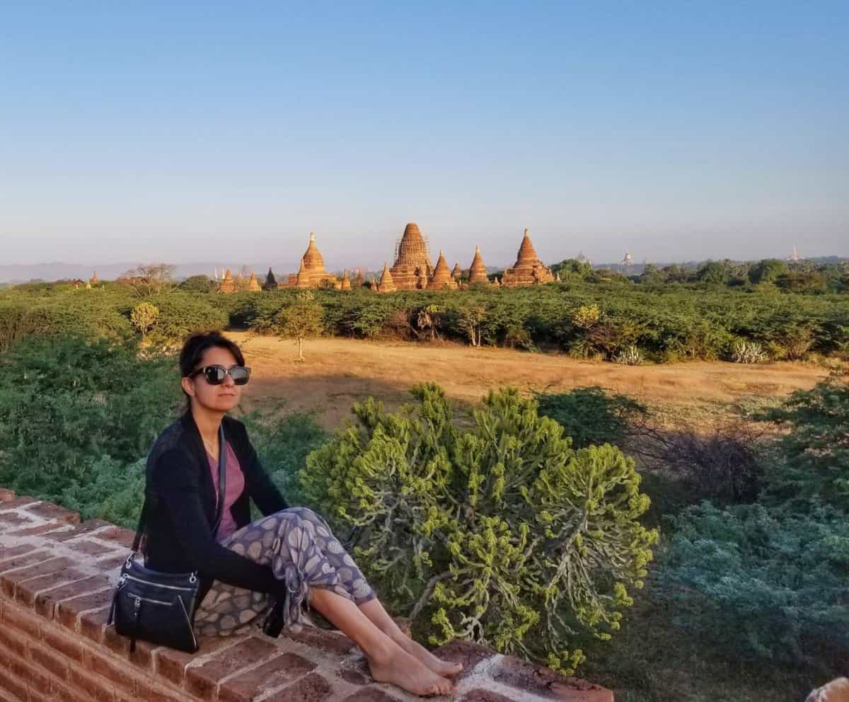 Bagan Burma Guide A person wearing sunglasses and casual clothing sits on a brick ledge, with a backdrop of ancient temples and green foliage in Bagan, Myanmar.