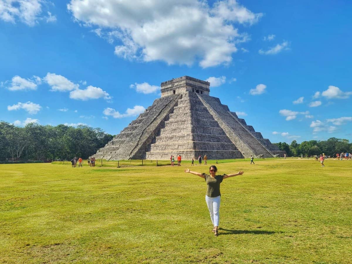 A woman standing with her arms outstretched in front of the Kukulkan Pyramid in Chichen Itza, Yucatan, Mexico. The pyramid is made of stone and has a steep incline. The sky is blue and there are some clouds in the background.