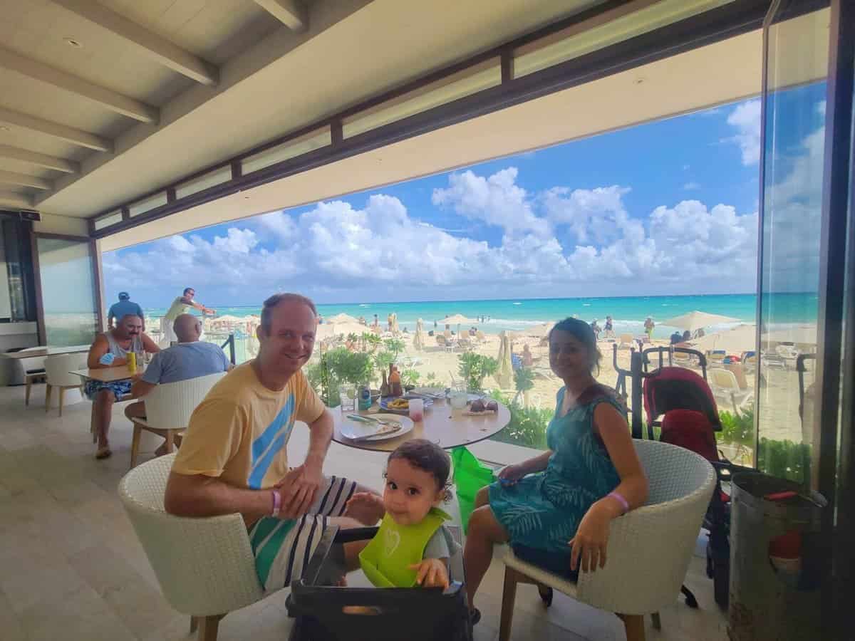 A family seated at a table indoors at Mamitas Beach Club in Playa del Carmen, with a stunning view of the beach and turquoise sea in the background.