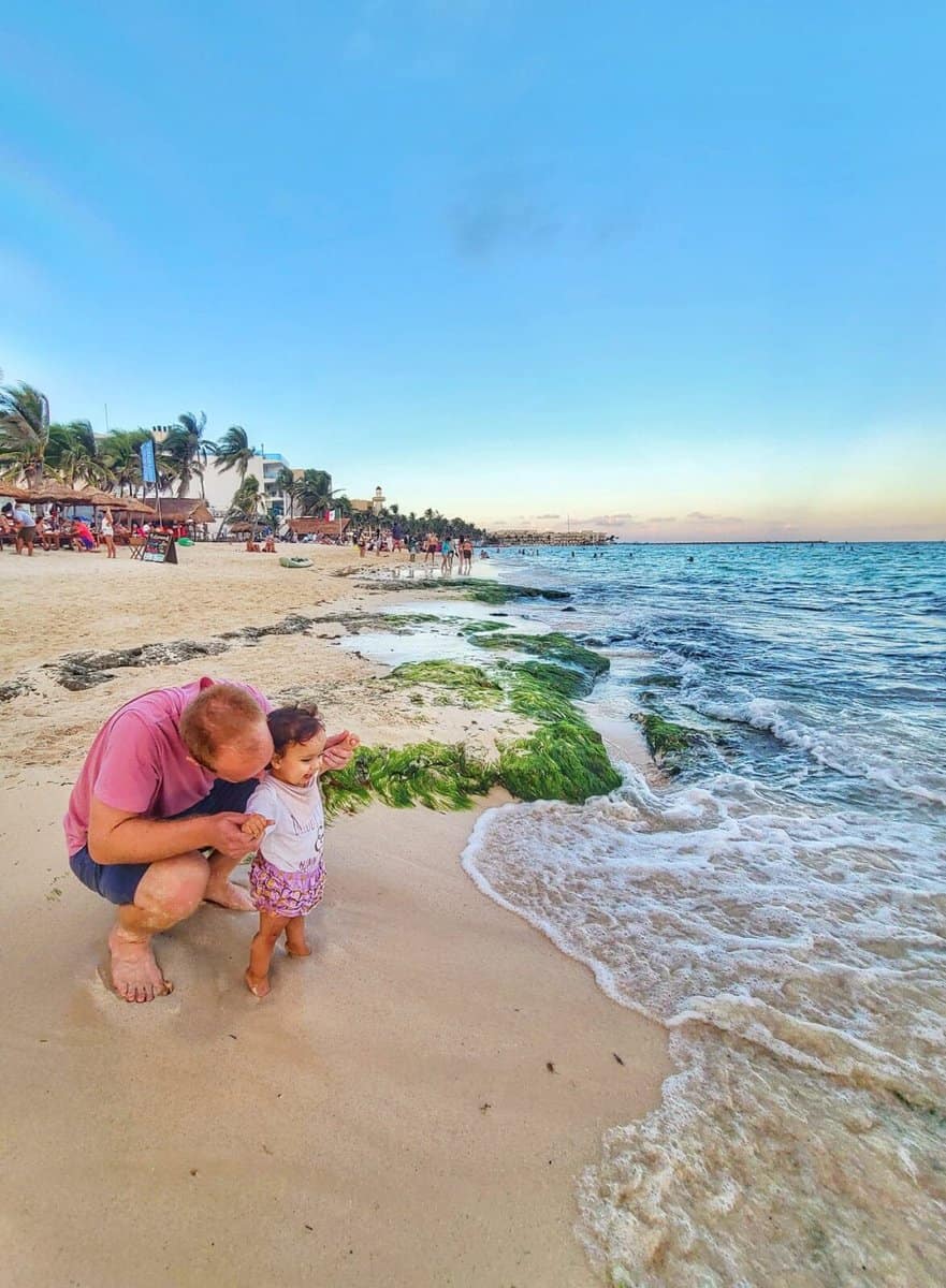 A man helps a toddler walk along the sandy beach near the water's edge at Playa del Carmen, Mexico.