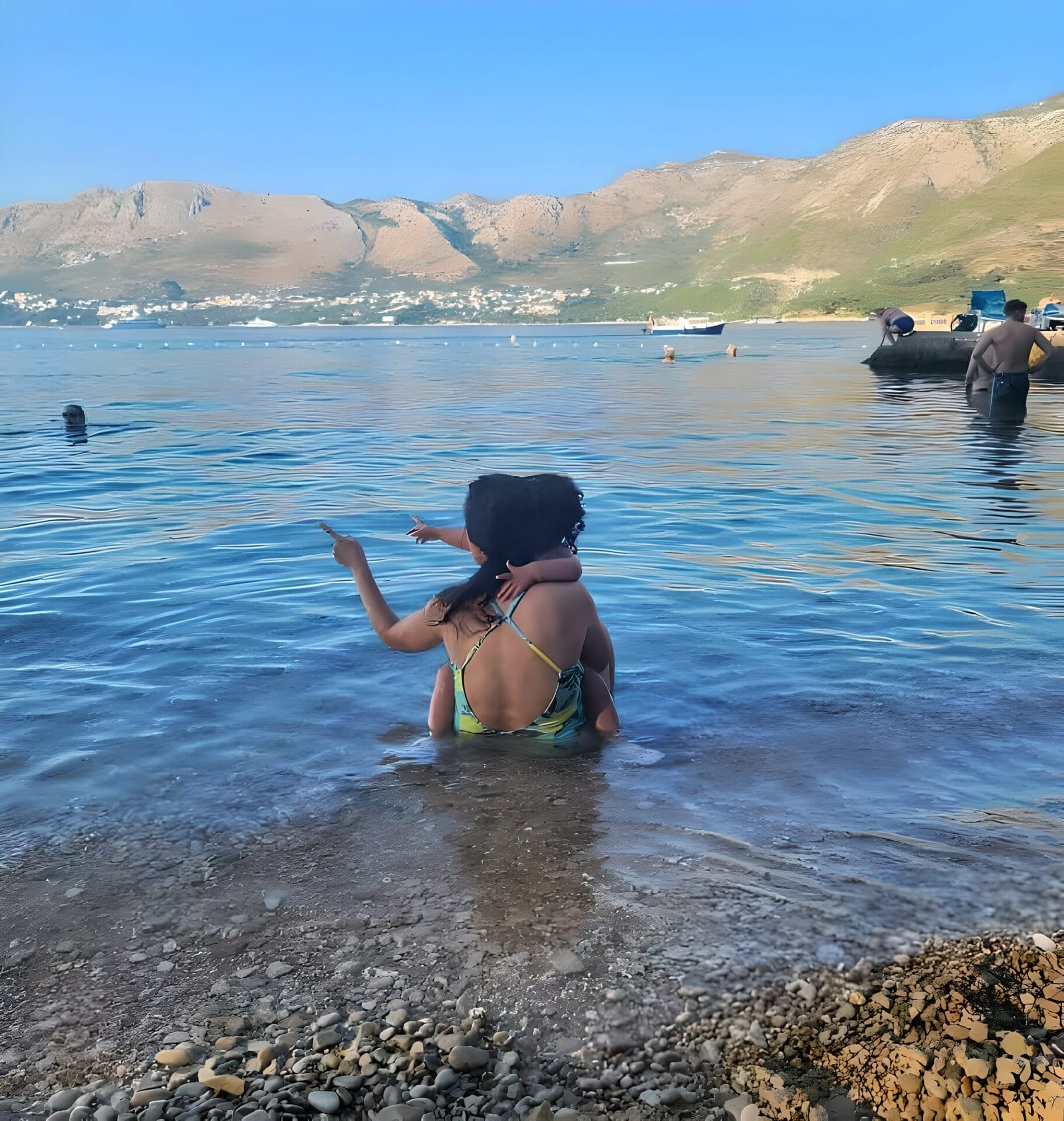 A woman in a swimsuit sits on a rocky shore in Cavtat, Croatia, holding a child. They are both looking out and pointing towards the water, surrounded by mountains and a partially clear blue sky in the background.