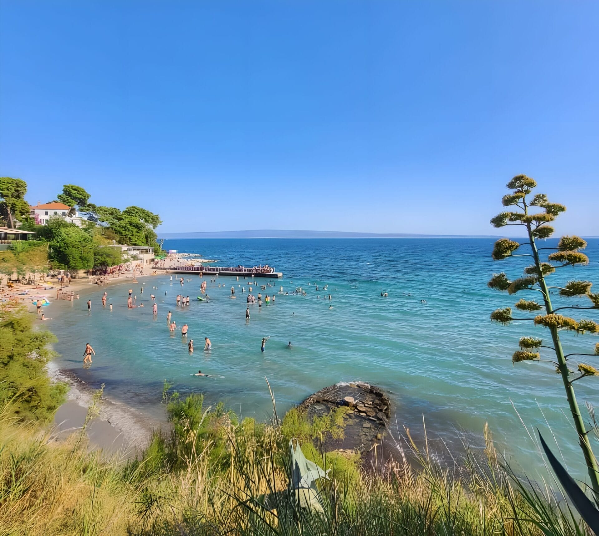 Family Beach in Split, Croatia A scenic view of a crowded beach in Split, Croatia, with clear blue water and people swimming. The coastline is dotted with greenery and a small pier extends into the sea. The sky is clear and bright blue.