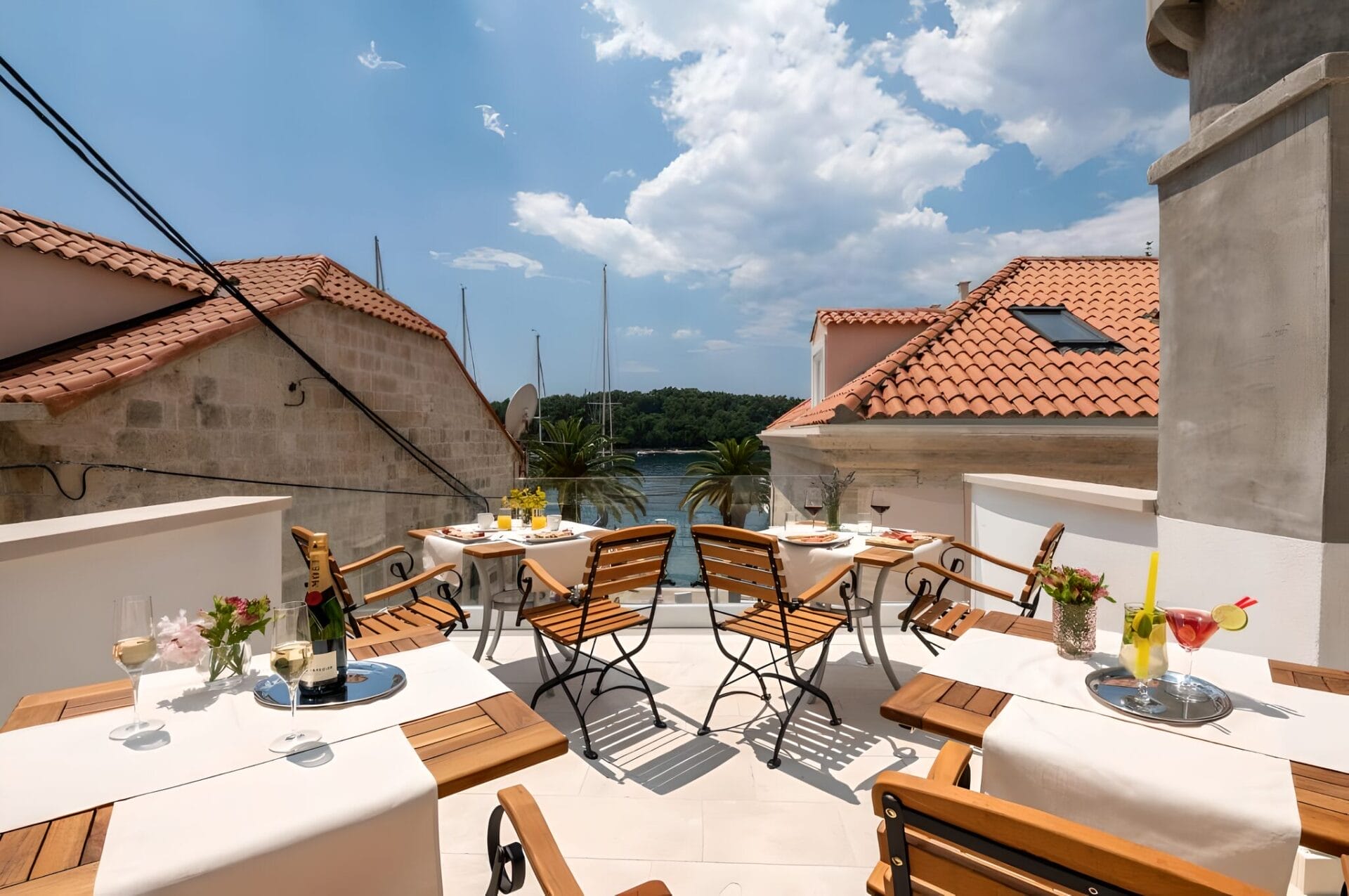 Outdoor terrace dining area at Boutique B&B Villa Faggioni in Cavtat, Croatia, featuring table settings with drinks and flowers, overlooking a view of rooftops and the sea.
