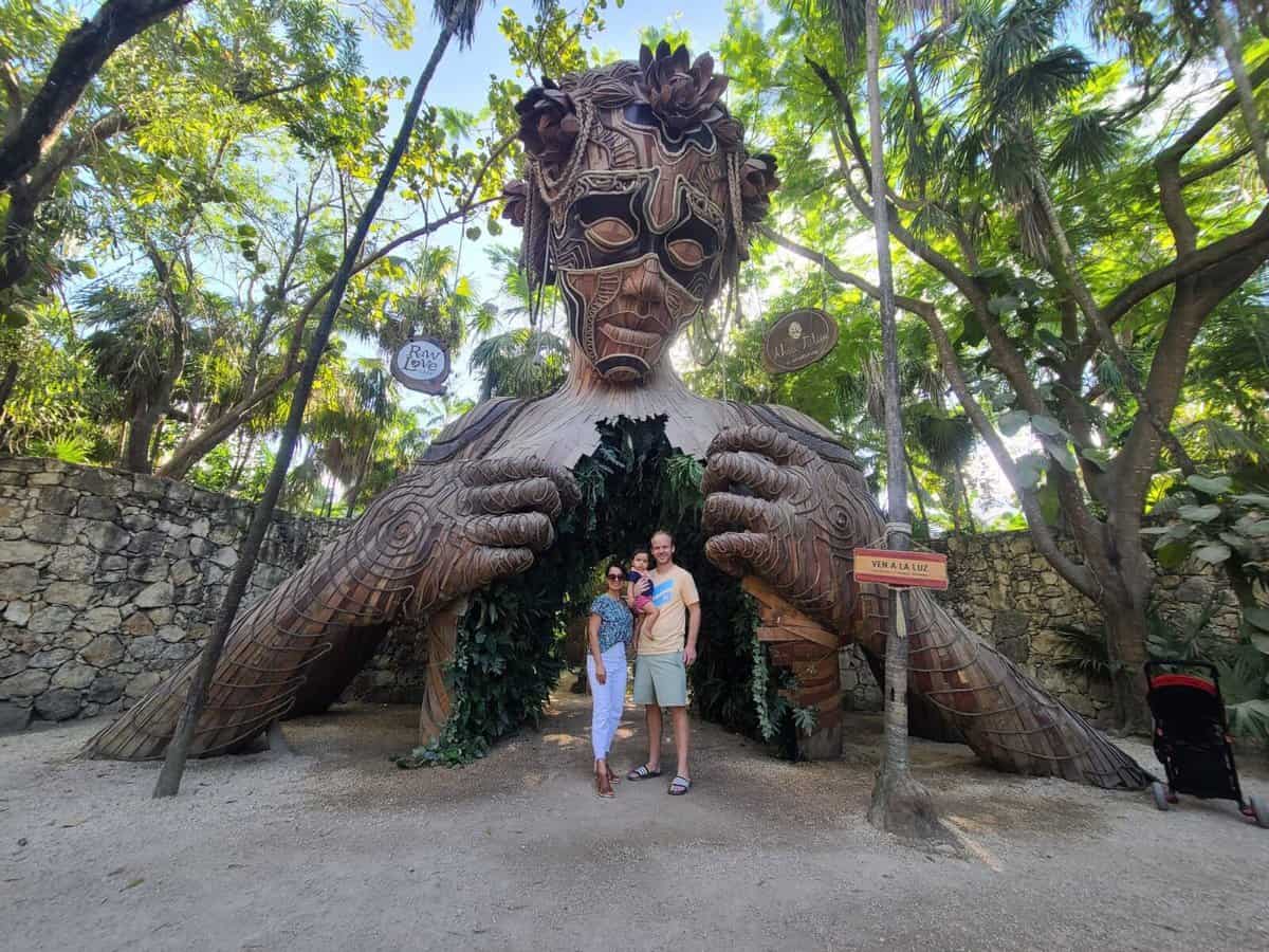 A couple stands in front of the "Ven a la Luz" sculpture in Tulum, Mexico, an intricate wooden structure depicting a large figure with open arms, set in a lush, green area with a stone wall in the background.