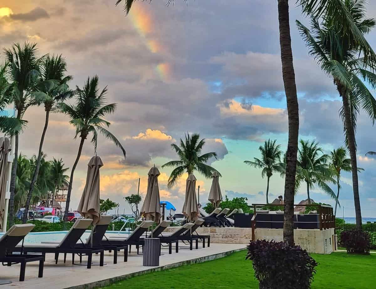 Poolside loungers with umbrellas surrounded by palm trees under a cloudy sky with a rainbow in Playa del Carmen, Quintana Roo, Mexico.